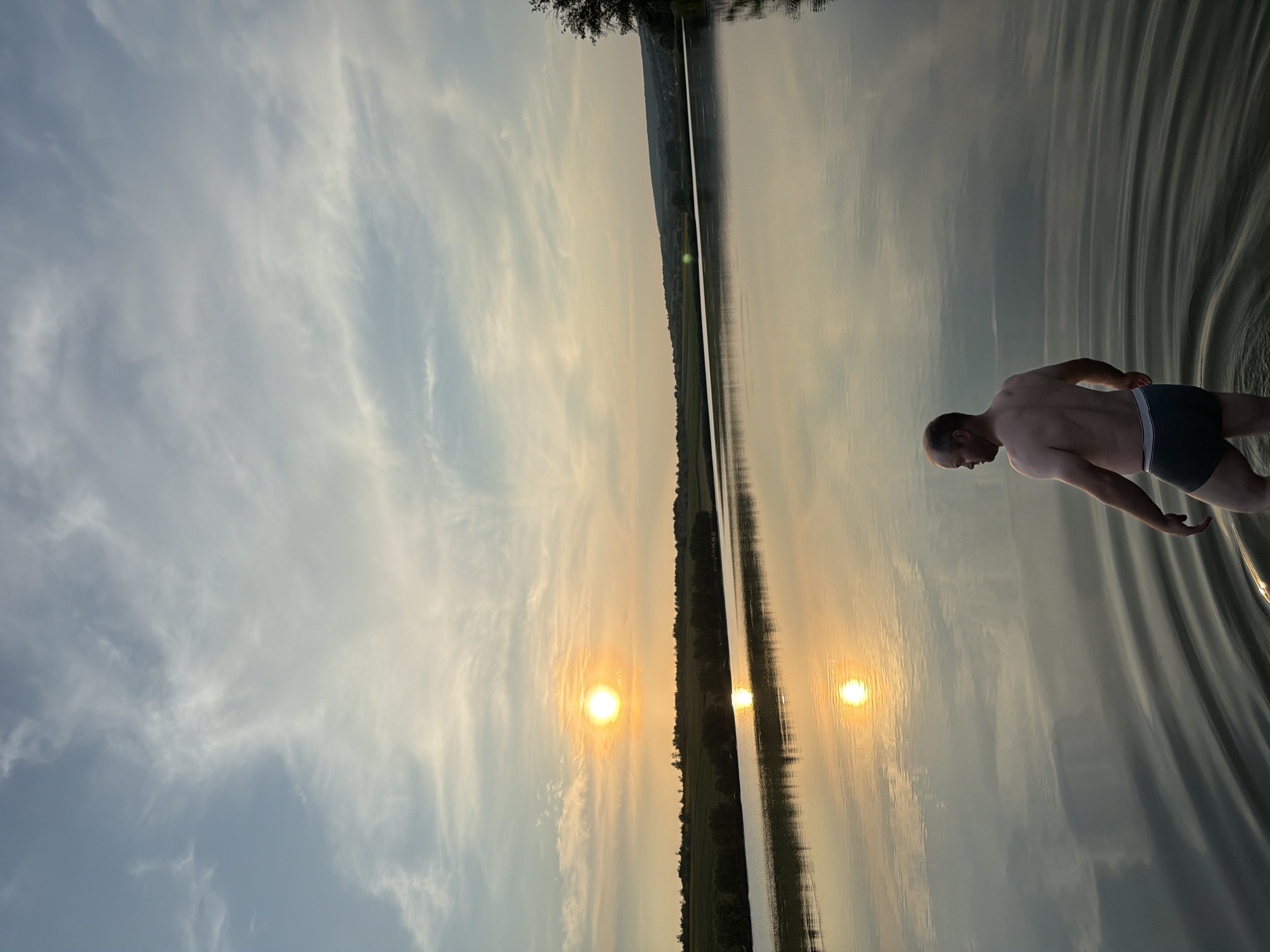 An outdoor, vertical, medium shot captures a man standing in the shallow water of a lake at sunset. The man, facing away from the camera and to the left, has fair skin, a shaved head, and appears to be wearing dark swim trunks. He is looking out towards the horizon. The lake's surface is calm, with concentric ripples radiating outwards from where the man is standing, suggesting movement through the water. The sky is a gradient of pale blue and soft orange hues, with wispy clouds stretching across. The sun, a bright, glowing orb, is positioned in the upper left quadrant of the frame, casting a warm, golden reflection on the water. A tree line, punctuated by fields and some vegetation, forms a distant horizon. The scene appears tranquil and natural, suggesting a rural or semi-rural setting, possibly in Găleștii Noi, Moldova, given the context.