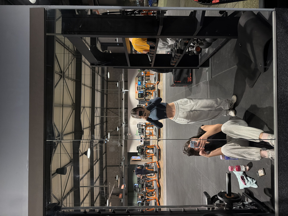 The mirror reflects a gymnasium interior with two young women posing. One stands, facing the mirror and holding a phone. The other sits with gym accessories nearby. Behind them are treadmills and various workout machines. The ceiling is exposed, with a metal frame and industrial lighting. Above them in the reflection is a weight rack with dumbbells. The lighting in the gym is bright, highlighting the orange accents of the equipment.