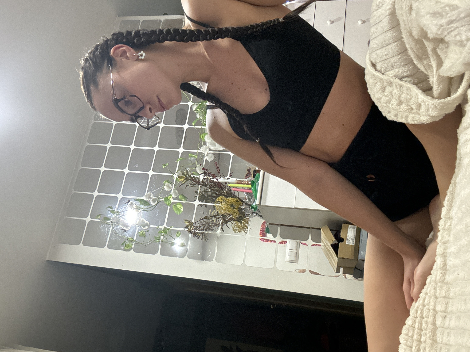 An indoor, vertical shot shows a young woman in what appears to be a bedroom or living space in Krasnoyarsk, Russia. She is turned slightly to her left, with her head tilted downwards, gazing off-frame. Her dark hair is styled in two prominent braids falling over her shoulders. She wears dark-rimmed glasses and a white, daisy-shaped earring in her left ear. Her attire consists of a black sports bra or crop top and black shorts. Her skin is fair, with several small freckles or moles visible on her torso.
Her hands are positioned near her upper thighs, partially obscured by a white, textured blanket or towel that occupies the right side of the frame. Her expression is calm and introspective.
The background features a white modular unit, possibly a wall panel or shelving, with square sections, some of which are reflective. Green vines with small white flowers are draped across these panels, and a dried floral arrangement sits atop the unit. Other items like small boxes and a light switch are also visible. The lighting is artificial and appears dim, suggesting it is evening or night. The lower background is dark, possibly indicating a television screen or dark furniture. No discernible text is visible in the image.