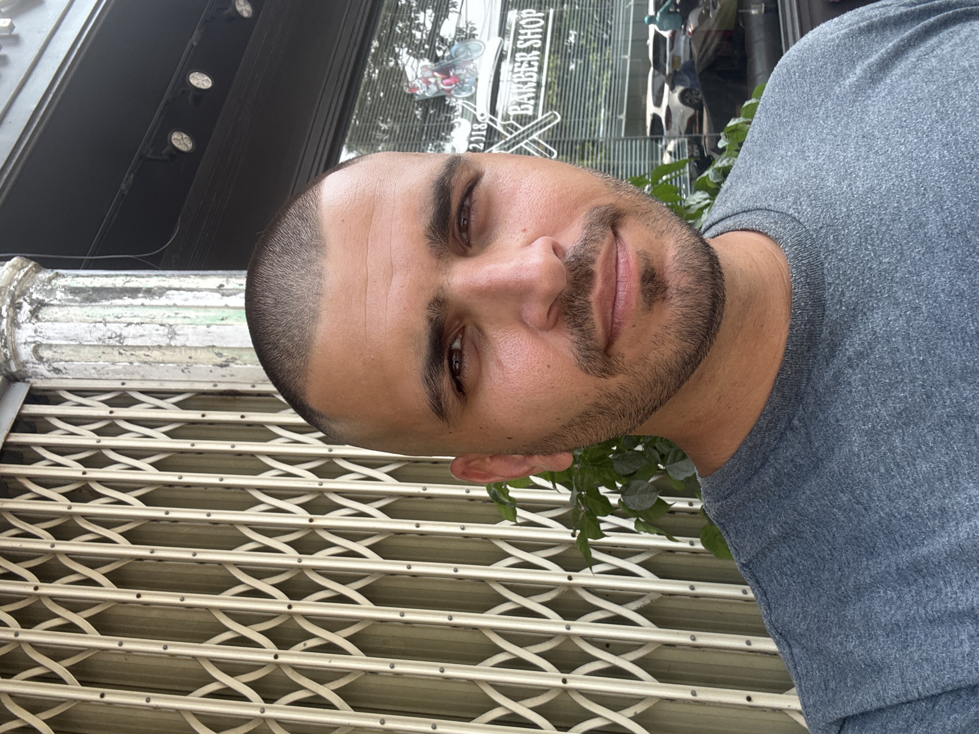 A man stands in front of a building. The sign on the building reads "BARBER SHOP." The man has short, dark hair and a beard. He is wearing a gray t-shirt. Behind him is a metal security gate and a column. The location is Đà Nẵng, Vietnam.
