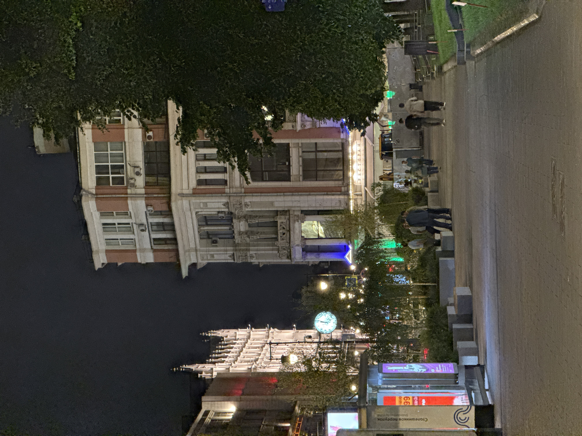 This image depicts a night scene on a city street, likely in Moscow, Russia, given the presence of Cyrillic text on signage. In the foreground, a paved walkway stretches into the distance, flanked by planters and grass areas. Several trees are visible, some illuminated with decorative lights.
On the left side of the street, a modern building with illuminated signage and displays is present. Further down the street, a tall, ornate building with Gothic architectural elements is brightly lit. Across from this, another building displays a sign that reads "РЕСТОРАН" (Restaurant).
Several individuals are walking along the street. Two men are walking away from the camera in the middle ground. A group of people is standing and walking in the middle distance, near the trees and planters.
The overall atmosphere is that of a bustling urban environment at night, with a mix of historical and modern architecture, commercial activity, and pedestrian traffic.