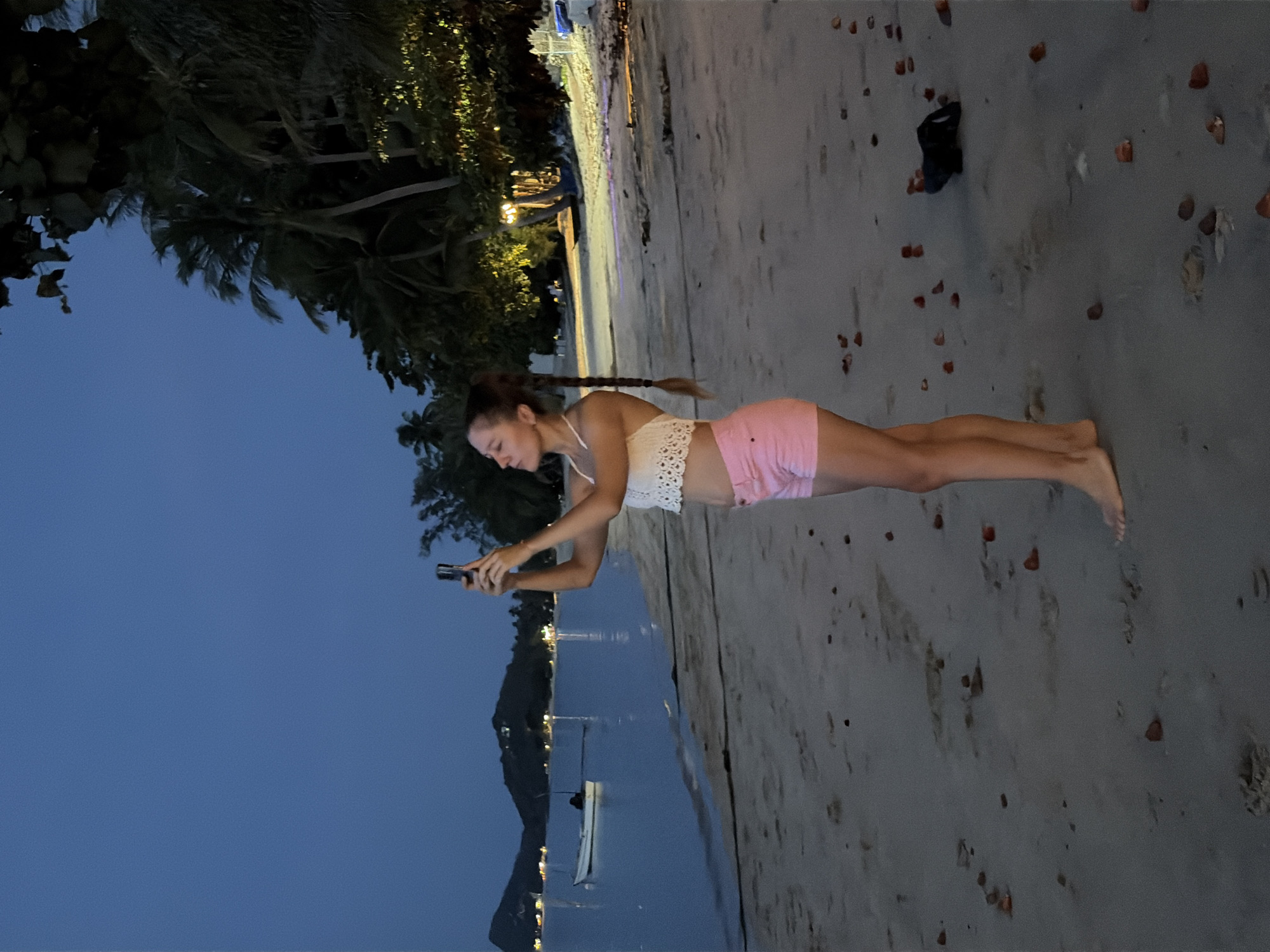 A young woman with a braided ponytail stands on a sandy beach at dusk, holding a smartphone. She is wearing a white lace crop top and pink shorts. The beach is lined with palm trees and illuminated by artificial lights in the distance. The ocean is calm, with a few boats visible on the water. The sky is a deep blue, indicating the time of day is twilight. The overall mood is peaceful and serene, with the woman appearing relaxed as she uses her phone. There is no visible text in the image. The setting is likely a tropical beach resort in Baan Thong Po, Thailand, given the presence of palm trees and the serene atmosphere.