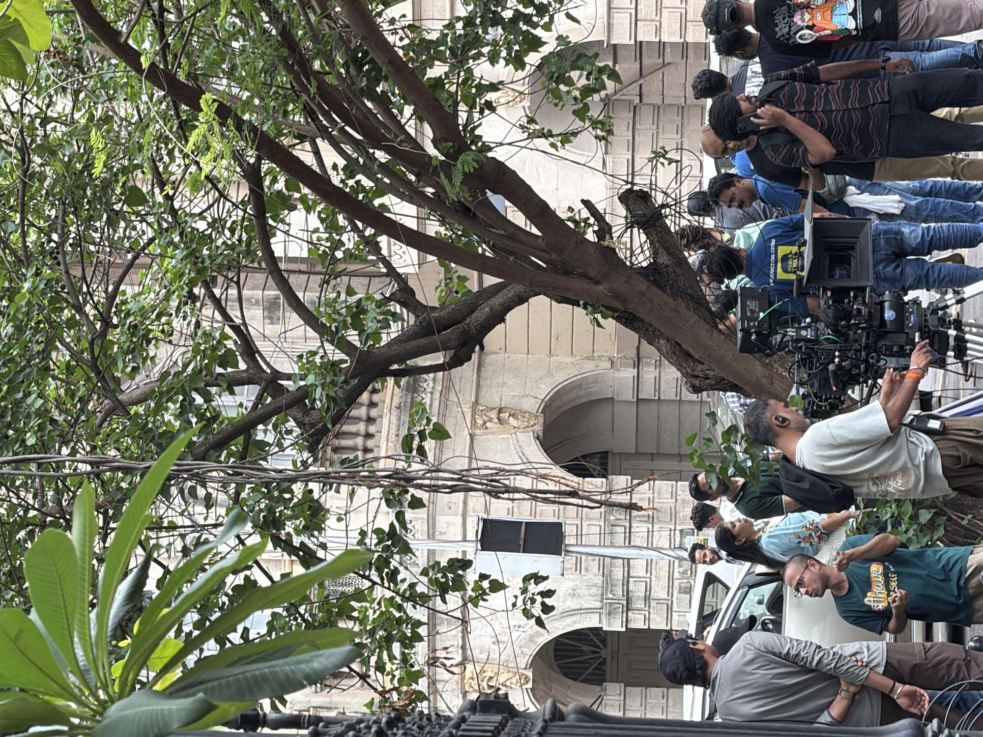 The image depicts a group of people gathered on a street in Mumbai, India, likely a film or television production site given the presence of professional camera equipment, including a large cinema camera on a tripod and a director's chair with a visible "PRODUCTION CREW" label. A mature tree with a thick trunk and branches shades a portion of the scene, with its leaves and hanging vines obscuring parts of the background.
In the foreground, a person in a green t-shirt with the word "have" visible is gesturing with their hands, appearing to be in conversation. To their left, another individual wearing a grey long-sleeved shirt and brown pants stands with their back to the camera. A large white vehicle is parked behind them.
The background features an old, ornate building with arched entrances and decorative architectural elements, characteristic of colonial-era structures found in Mumbai. The building is constructed of light-colored stone or brick. Several individuals are seen milling around, some looking towards the camera, others engaged in conversation. One person is on the phone, and another is wearing headphones, possibly a sound technician. The overall atmosphere suggests an active and organized setting, likely a busy urban environment.