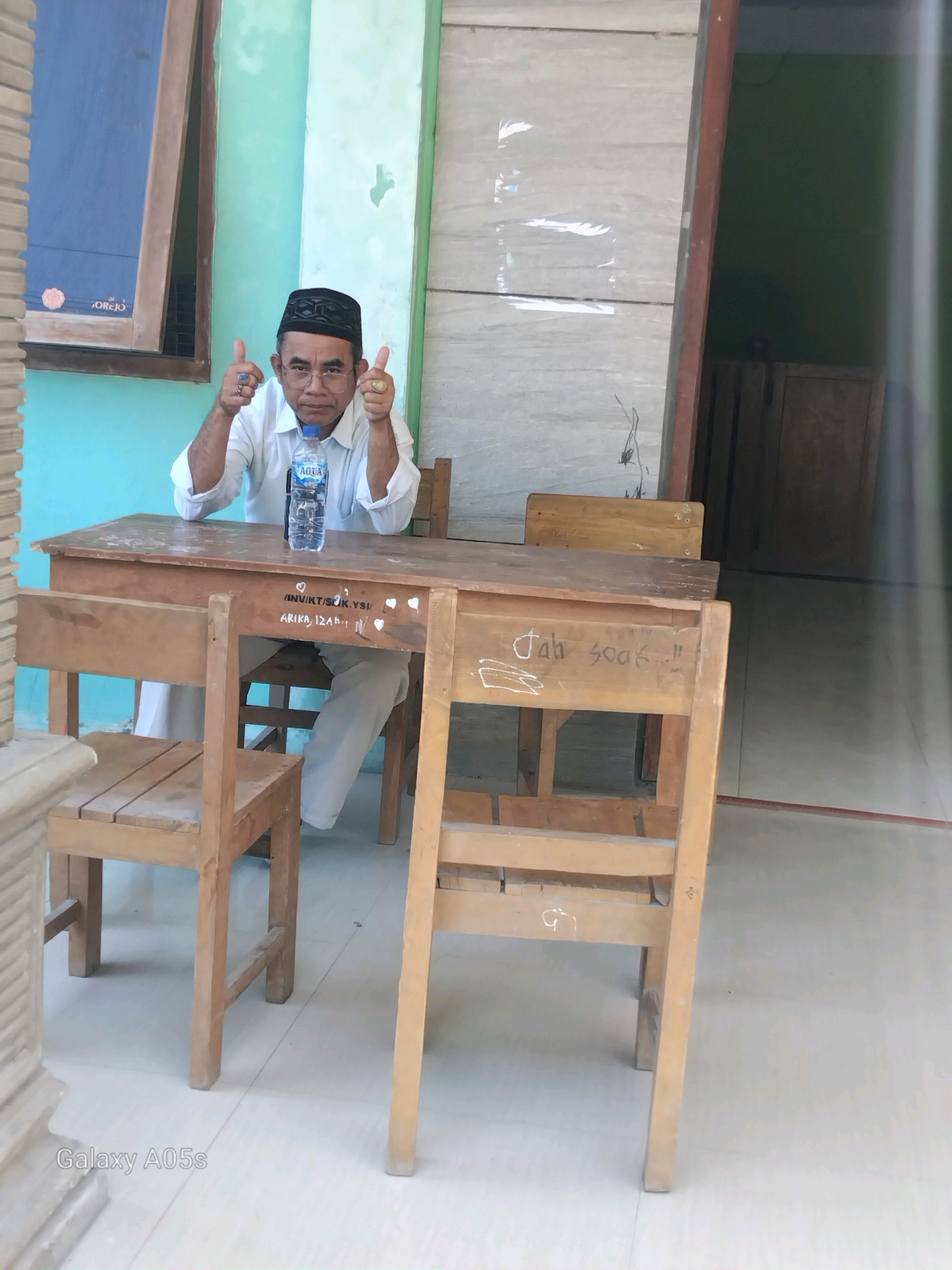 A man wearing a white long-sleeved shirt, white pants, and a black peci is sitting at a wooden desk in what appears to be a classroom in Karanganyung, Indonesia. He is giving a double thumbs-up with both hands, and a bottle of Aqua water sits on the desk in front of him. There are two wooden chairs in front of the desk, and another wooden chair behind the desk. The walls are painted light green and gray. The floor is tiled with white tiles. There is a window on the left with a blue pane. The man is wearing glasses and rings on his fingers. The desk has some writing on it, including "/INV/KT/SCK/YS/" and "ARIKA, 12A", and "Jah 3046!!". The image is watermarked with "Galaxy A05s".