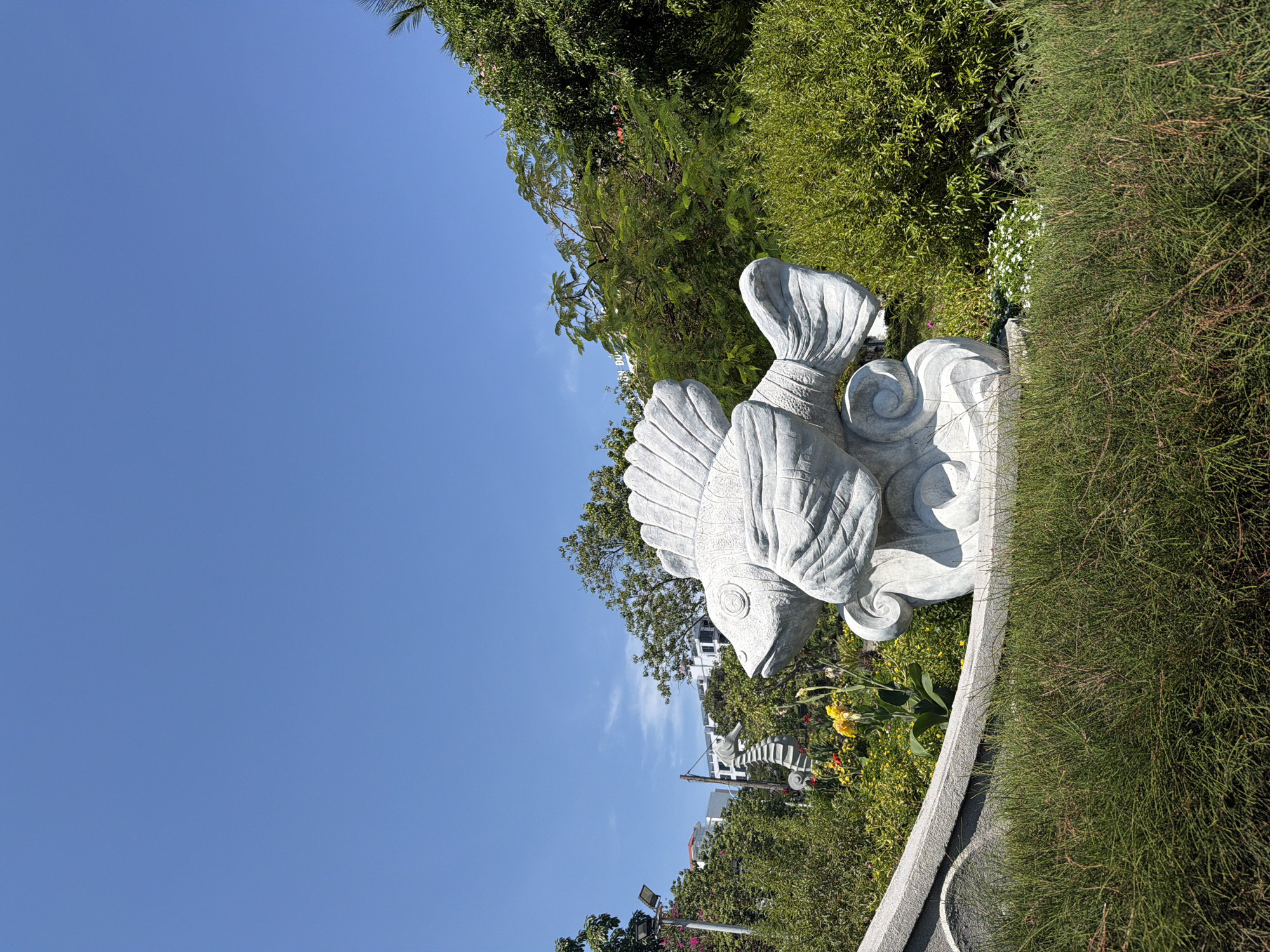 A fish statue, likely made of stone or concrete, is the focal point of the image. It is positioned on a low, curved wall. The statue appears to be a stylized representation of a fish with prominent fins and scales.
The statue is surrounded by vegetation including trees, bushes, and ground cover of various textures and shades of green.
The background is a clear, blue sky.
The setting is Bac Nha Trang Ward, Vietnam, based on contextual information. The presence of the fish statue suggests a park, garden, or public space. Buildings and other structures are visible in the background indicating that this is likely an urban area.