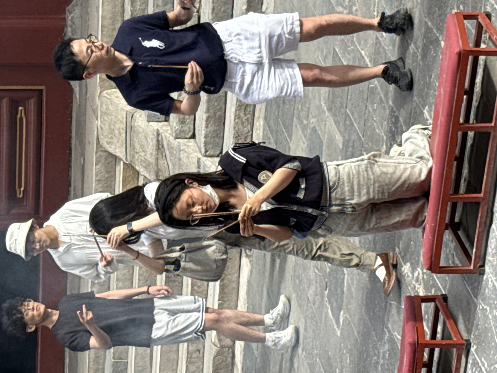 In Dongcheng District, China, on a paved courtyard with stone steps leading up to a red-lacquered building, four individuals are engaged in a ritual involving incense. In the foreground, a woman kneels on a red bench, holding a lit incense stick in each hand and a mask hanging around her neck. To her left, a man wearing a dark blue polo shirt and white shorts holds a single incense stick. Further left and behind him, a man in a white hooded jacket and a bucket hat also holds an incense stick. On the far left, a man in a black t-shirt and grey shorts gestures with his left hand. The scene suggests a religious or ceremonial observance.