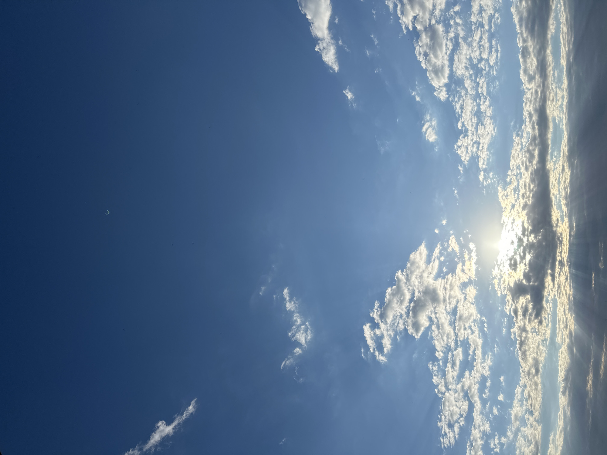 The image shows a wide expanse of blue sky with scattered clouds. The sun is visible, partially obscured by clouds, casting light rays downward. The clouds are a mix of bright white and darker, shadowed forms. A crescent moon is faintly visible in the upper portion of the sky. There is no discernible urban landscape or activity present in the image, suggesting an open view, likely from a high vantage point or a location with minimal obstructions. The scene is natural and devoid of human presence or man-made structures.