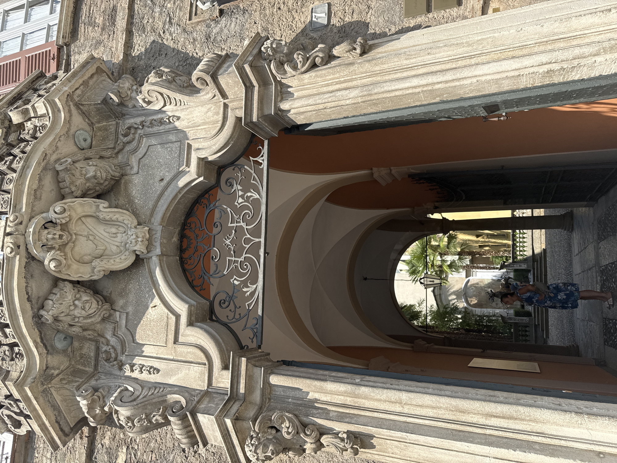 This is a low-angle, eye-level, outdoor, full shot of the ornate Baroque entrance archway to a building, likely in Lugano, Switzerland. The archway is made of stone and features intricate carvings of faces and decorative elements. In the center of the archway is a wrought iron gate. Beyond the archway, an arcaded walkway leads to a courtyard. A woman in a blue floral dress is standing in the walkway, looking at a plaque on the wall. Sunlight casts shadows on the building and walkway. The overall impression is one of grandeur and history.