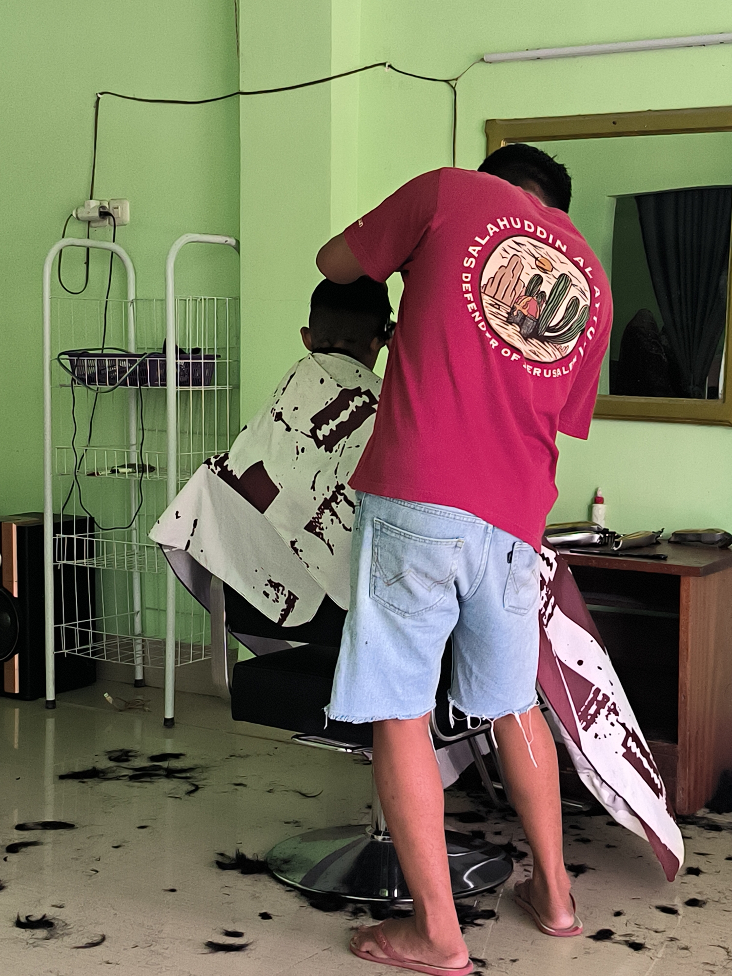 In Bumi Mulyo, Indonesia, a barber is cutting a child's hair. The barber, wearing a red t-shirt and denim shorts, stands behind the child who is seated and draped with a cape. The room is painted green and the floor is littered with hair clippings. A mirror hangs on the wall, reflecting a window with dark curtains. Shelving units hold various items, and electrical cords are visible. A small wooden cabinet with tools is also present.