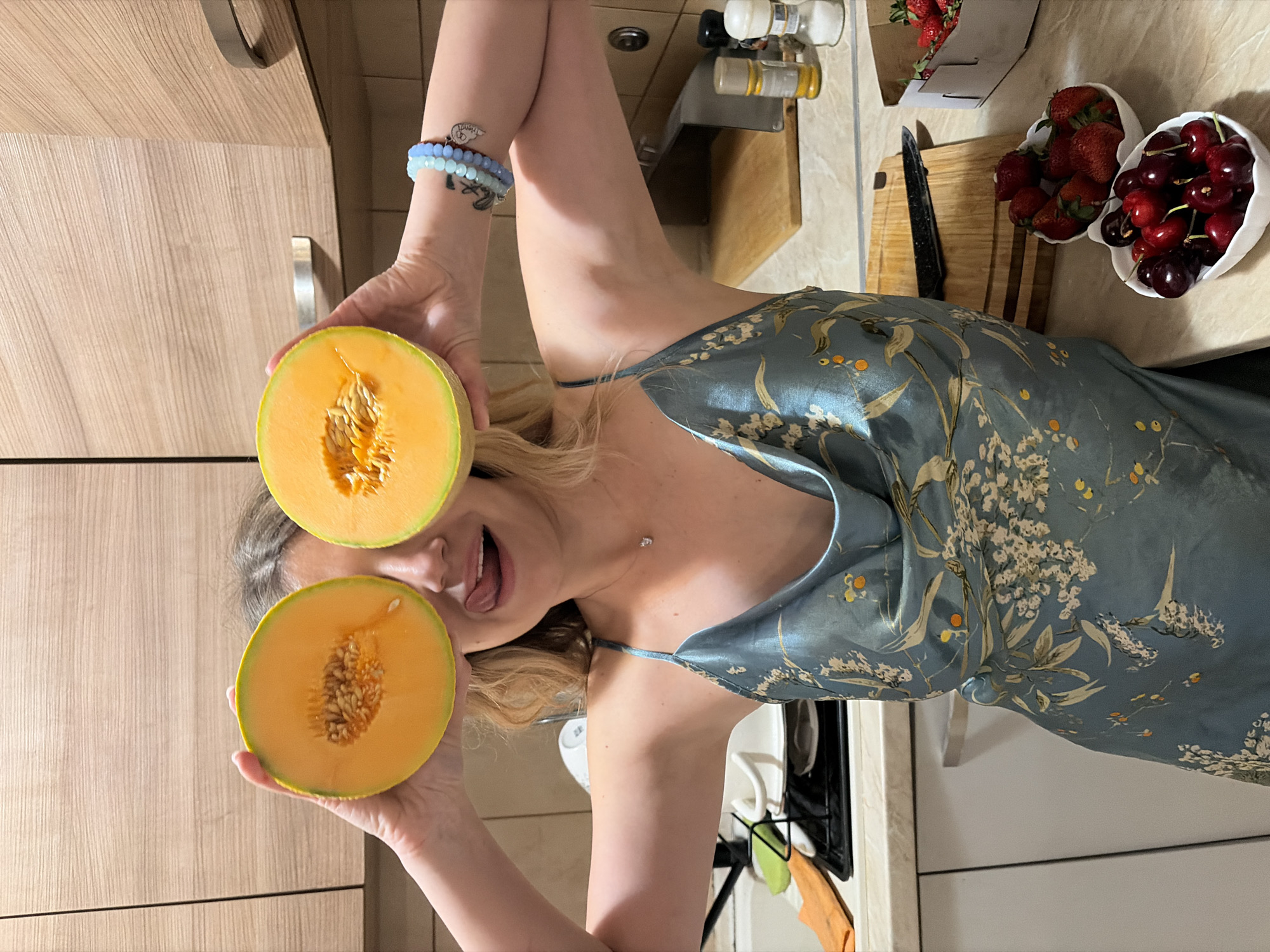 A woman is in a kitchen, holding two halves of a cantaloupe in front of her eyes. She is wearing a blue floral patterned dress and several beaded bracelets on her left wrist. Her mouth is open, and her tongue is sticking out. In the background, there is a counter with a wooden cutting board, a black knife, strawberries in a cardboard container, and two white bowls filled with strawberries and cherries. The kitchen appears to have wooden cabinets. The location cannot be determined as being in Budva, Montenegro, based on the visual information alone.