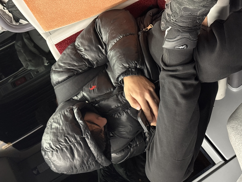 A person sits on a train, wearing a black puffer jacket with the hood up and black tracksuit pants. Their hand holds a smartphone. A pair of grey trainers rests on the jacket. The train's interior features orange seats, black paneling, and white accents. The overall lighting is muted, creating a casual, everyday scene.