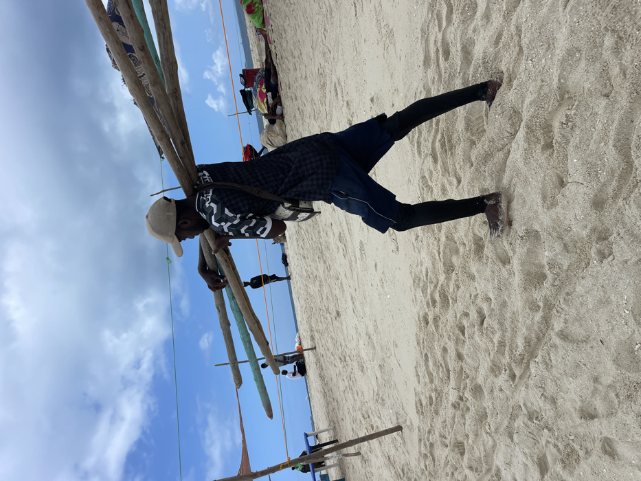 The image displays a sandy beach scene in the city of Zanzibar, Tanzania. A male individual, positioned in the foreground and facing right, is depicted carrying multiple long wooden poles horizontally across his left shoulder and back, stabilizing them with both hands. He wears a light-colored baseball cap, a dark patterned short-sleeved shirt, dark blue shorts over black leggings, and dark footwear. A light-colored shoulder bag with a dark strap hangs from his right side. The ground consists of light-colored sand, marked with footprints. In the middle ground, a dark-clad individual stands upright further left, near the water's edge. To the far left, two other individuals are seated near a light-colored table-like structure. To the right of the primary subject, multiple individuals are resting or lying on the sand, some covered by colorful fabrics. Several vertical wooden poles with horizontal lines (ropes or wires) stretched between them form a visible structure or boundary across the mid-ground. The sky above is partly cloudy with significant blue areas, and the ocean is visible as a distant horizon line.