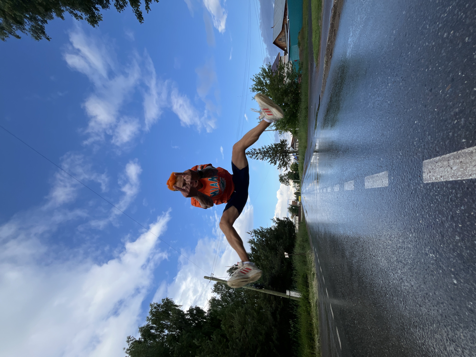 A young man with bright orange hair is captured mid-air performing a high jump with his legs spread wide in a split. He is wearing an orange t-shirt with the word "MIAMI" and a dolphin graphic, along with dark blue shorts and white athletic shoes with red stripes. He is on a wet asphalt road with visible white dashed lines, suggesting it may have rained recently. The road stretches into the distance, flanked by green trees and grass on either side. In the background, there are houses with wooden roofs, indicating a rural or suburban setting, likely in Chaikovsky, Russia. The sky is blue with scattered white clouds. The perspective is low-angle, emphasizing the man's airborne action and the vastness of the sky.