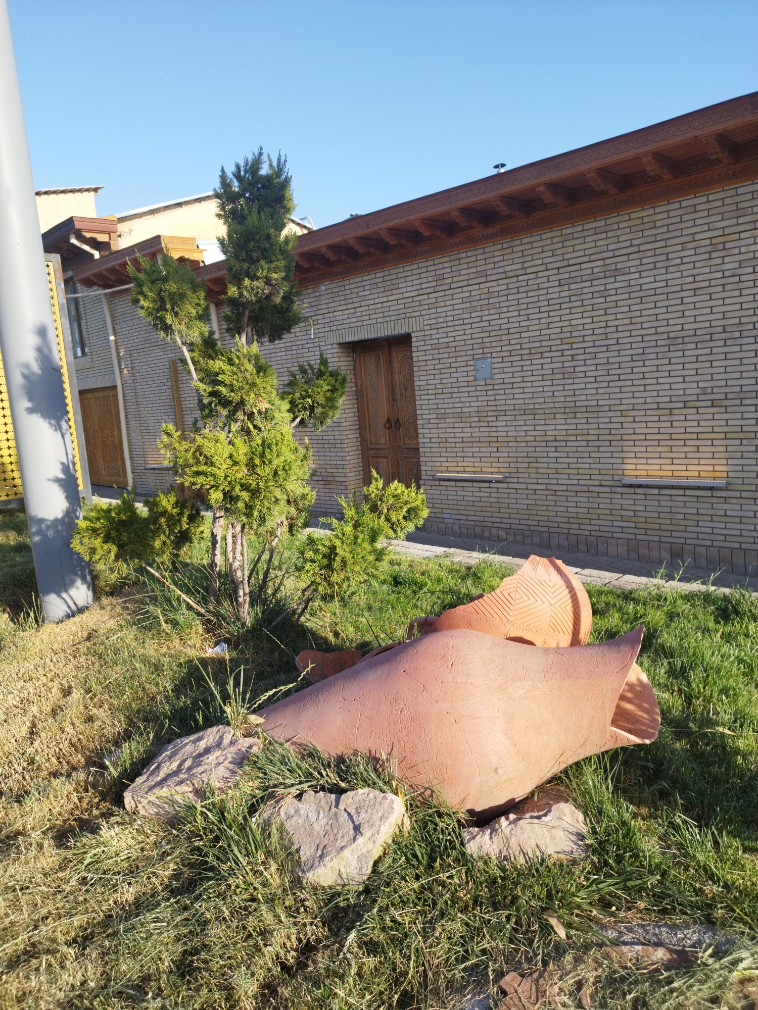 A low-angle outdoor shot shows a broken terracotta jug lying on the grass in front of a brick building. The jug is large and appears to be antique, with intricate patterns carved into its surface. Two small evergreen trees stand to the left of the jug, casting shadows on the building. The building is constructed of light-colored bricks and has a wooden roof with decorative trim. A gray pole stands to the far left of the frame, partially obscuring the building. The sky is clear and blue.