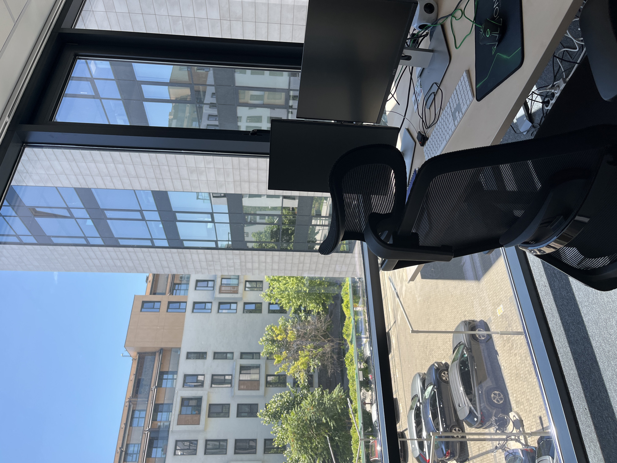 The image shows a modern office interior with a large window offering a view of the city. Outside, there are contemporary buildings, trees, and parked cars on a paved street. The office features a desk with two computer monitors, a keyboard, and a mouse. An office chair is positioned in front of the desk. The overall scene depicts a well-lit workspace with an urban backdrop. The architectural style of the buildings suggests a contemporary urban setting. The presence of multiple modern buildings, a paved street, and parked cars indicates an urban environment, likely within a commercial or residential district. The clear blue sky and the quality of light suggest it is daytime. The office setup with dual monitors and a mesh-back chair points to a professional or tech-oriented workspace. The image was taken from inside the office, looking out. The location is Plovdiv, Bulgaria.