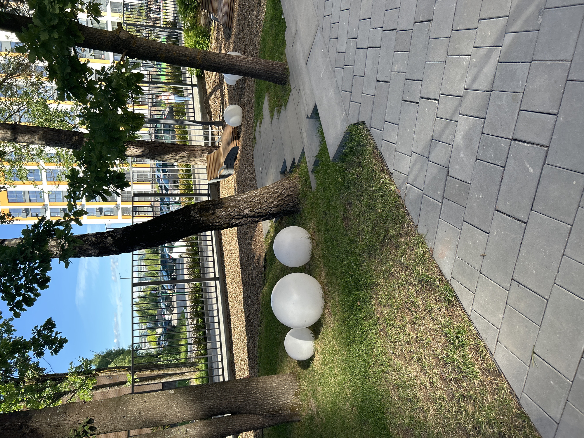 The image shows an outdoor scene with a modern urban aesthetic. In the foreground, a paved walkway made of rectangular grey stones runs diagonally across the frame. To the left of the walkway is a patch of green grass, where three white spherical objects, likely decorative lights or orbs, are placed. Two are larger and one is smaller, arranged in a staggered line.
Several trees with thick, textured bark are visible, with their trunks and branches extending into the frame. Some have lush green foliage. Behind the trees and grass, a black metal fence runs horizontally, separating the immediate area from a more distant scene.
Beyond the fence, a tall, modern building with a yellow and white facade stands prominently, featuring numerous windows. Parked cars and more greenery are visible in the middle ground. The sky is a clear, bright blue, suggesting a sunny day, likely in the late morning or early afternoon given the lighting. The overall impression is a well-maintained urban outdoor space, possibly a courtyard or park area within a residential complex. The location is identified as Kaluga, Russia. No people are explicitly visible, and no text is present in the image.