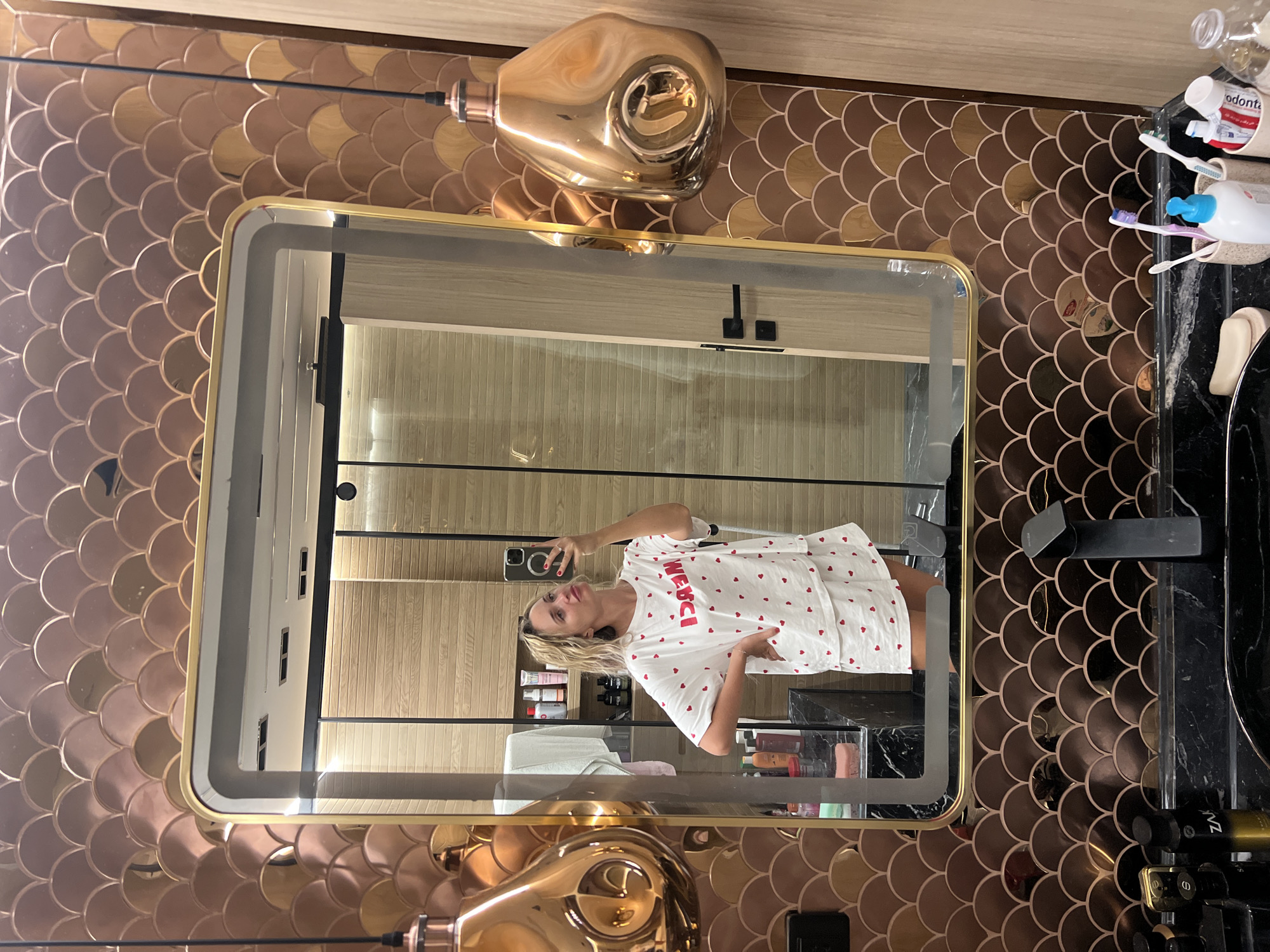 A woman is taking a mirror selfie in a bathroom. She is wearing a white pajama set with red hearts and the word "MERCI" printed on the top. The bathroom has a unique tiled wall with a scalloped pattern in shades of bronze and gold. A large mirror with a golden frame reflects the woman and the interior of the bathroom, which includes wooden paneled walls and a glass-enclosed shower. Two decorative copper lamps hang on either side of the mirror. On the counter, there are various toiletries, including bottles, toothbrushes, and a bar of soap. The woman is standing with one hand on her hip, holding her phone to take the picture. The overall aesthetic suggests a modern and stylish bathroom. This scene is located in Sharm El Sheikh, Egypt.