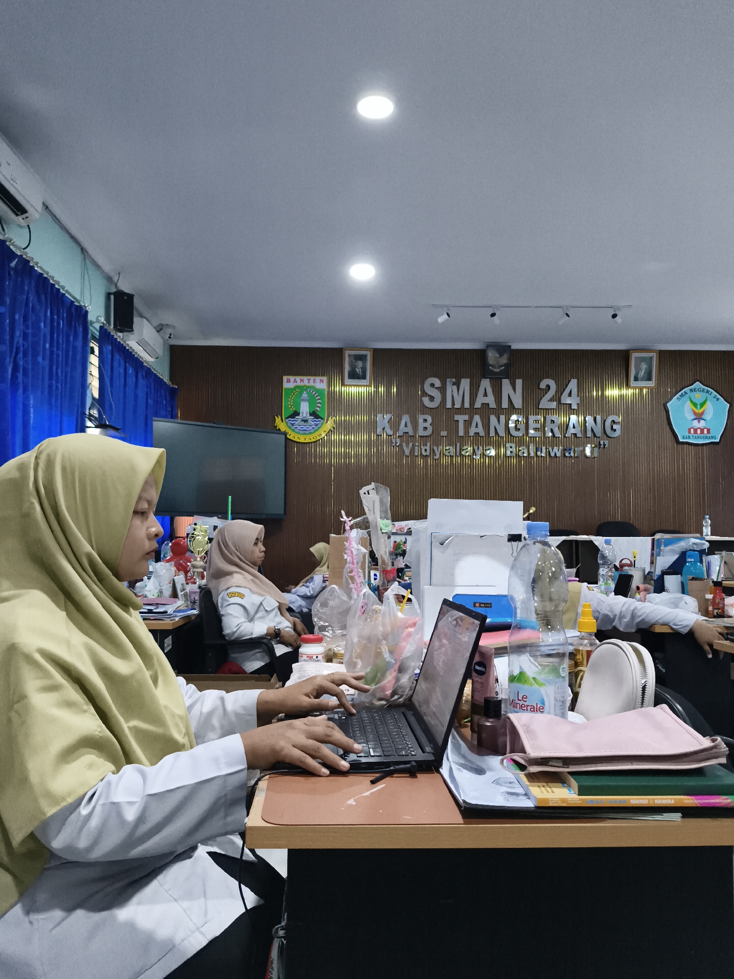 The image depicts an indoor scene, likely an office or classroom, at SMAN 24 Kab. Tangerang, Vidyalaya Bahuwarna, in Kuta Baru, Indonesia. In the foreground, a person wearing a light yellow hijab and a white uniform is seated at a desk, typing on a laptop. Their hands are visible on the keyboard. The desk is cluttered with various items, including a bottle of water labeled "Le Minerale," a pink pouch, books, and other office supplies. Behind this individual, another person in a similar uniform, also wearing a hijab, is seated and looking forward. In the background, a large screen is mounted on the wall, and the wall itself is decorated with a wooden paneling and signage that reads "SMAN 24 KAB. TANGERANG Vidyalaya Bahuwarna." There are also framed pictures and a school emblem on the wall. Blue curtains are visible on the left side of the frame. The overall environment appears to be a functional workspace with artificial lighting.