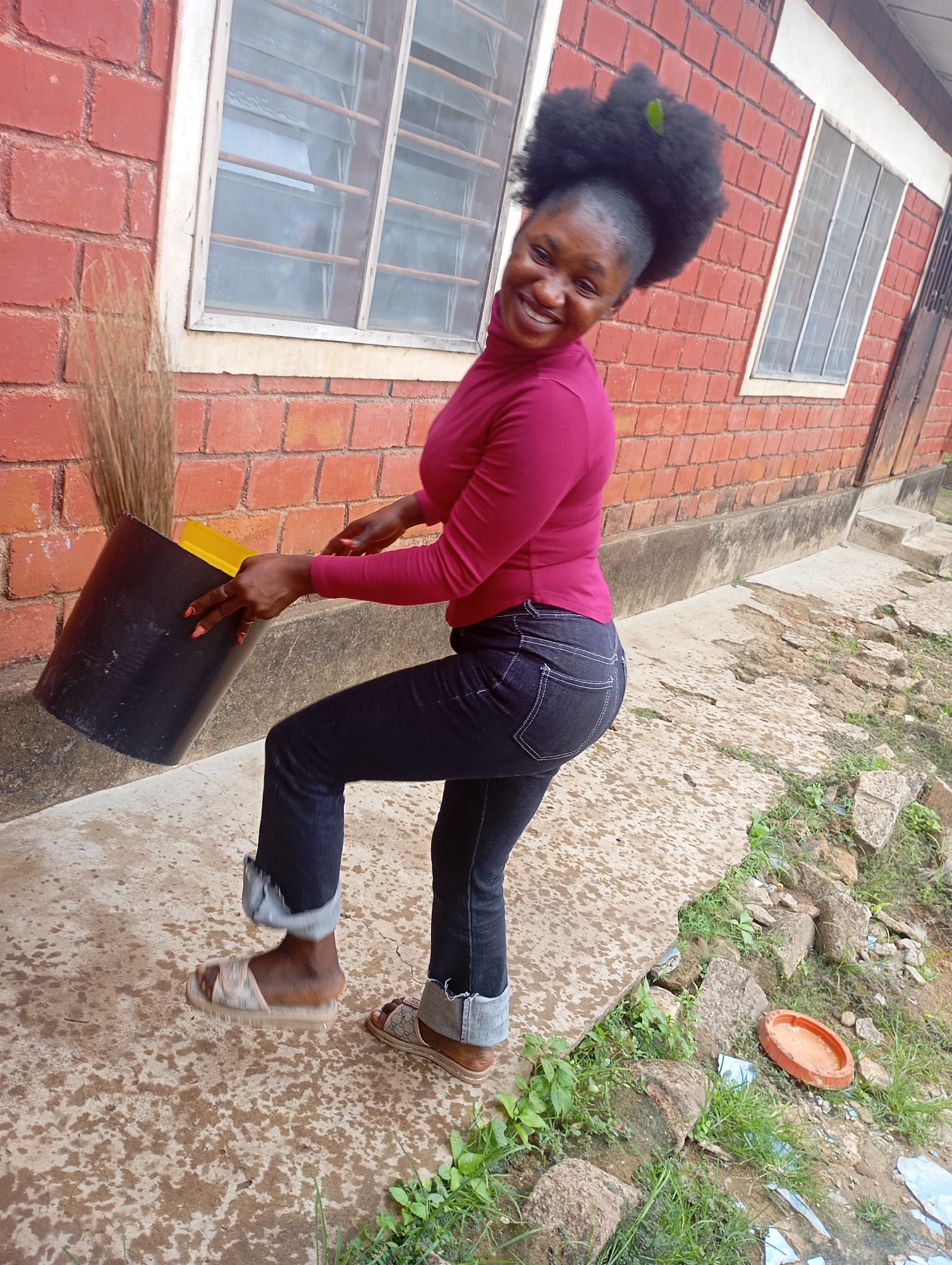 In New Stadium, Nigeria, a young woman with dark skin and voluminous curly hair is depicted. She is wearing a long-sleeved magenta turtleneck shirt and dark denim jeans, the cuffs of which are rolled up. Her feet are clad in beige slip-on sandals with a patterned sole.
The woman is actively engaged in an action, tilting a large black bucket that contains a yellow object, possibly a shovel or scooper, and a bundle of dry grass or straw, suggesting she is sweeping or cleaning. Her right hand is positioned on the rim of the bucket, while her left hand is near the top edge.
The background features a red brick building with white-trimmed windows. The ground is a mix of concrete and dirt, with scattered rocks and sparse vegetation. The scene appears to be outdoors, in a slightly neglected or working-class area, possibly a yard or alleyway adjacent to the building. The lighting indicates it is daytime.