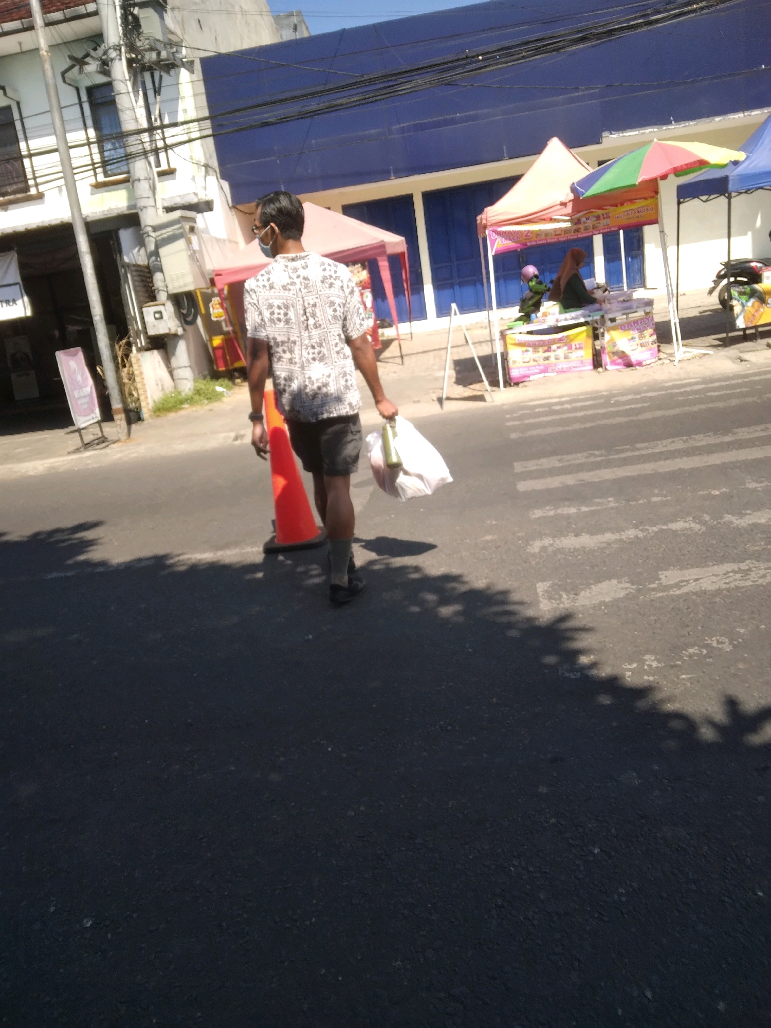A person wearing a patterned short-sleeved shirt, dark shorts, gray socks, and black shoes walks away from the camera, carrying a white plastic bag and a green bottle in their right hand. The person is walking on an asphalt road, past an orange traffic cone and a pedestrian crossing. In the background, there are tents set up with colorful awnings and various goods for sale. A building with blue doors and windows is visible behind the tents. Power lines are strung across the top of the image. The scene appears to be in a commercial area, likely a street market, in Sumampir, Indonesia, on a sunny day, as indicated by the shadows cast on the road.