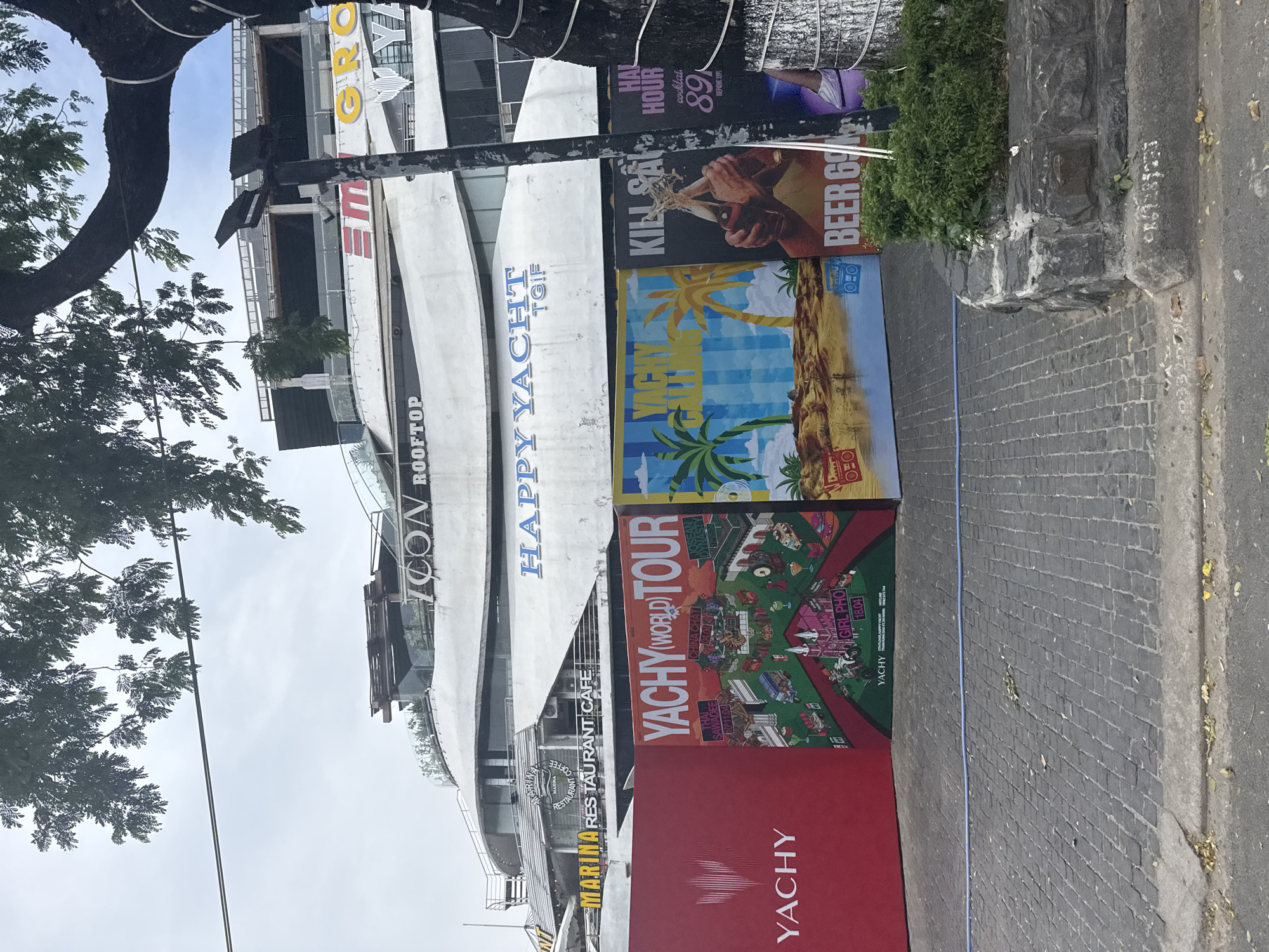 The image captures a street scene in Đà Nẵng, Vietnam, featuring a multi-story building with curved, white architectural elements. Signage indicates that the building houses "MARINA RESTAURANT CAFE" and "ICON ROOFTOP," along with "HAPPY YACHT TOIF." A large tree with dense foliage partially obscures the left side of the building. Posters attached to a street-side structure advertise "YACHY (WORLD) TOUR," "YACHY" itself, "KILL SA," "BEER 69," and "HAL HOUR cocktails 89." The foreground is composed of a brick-paved sidewalk with a street curb. The sky is overcast.