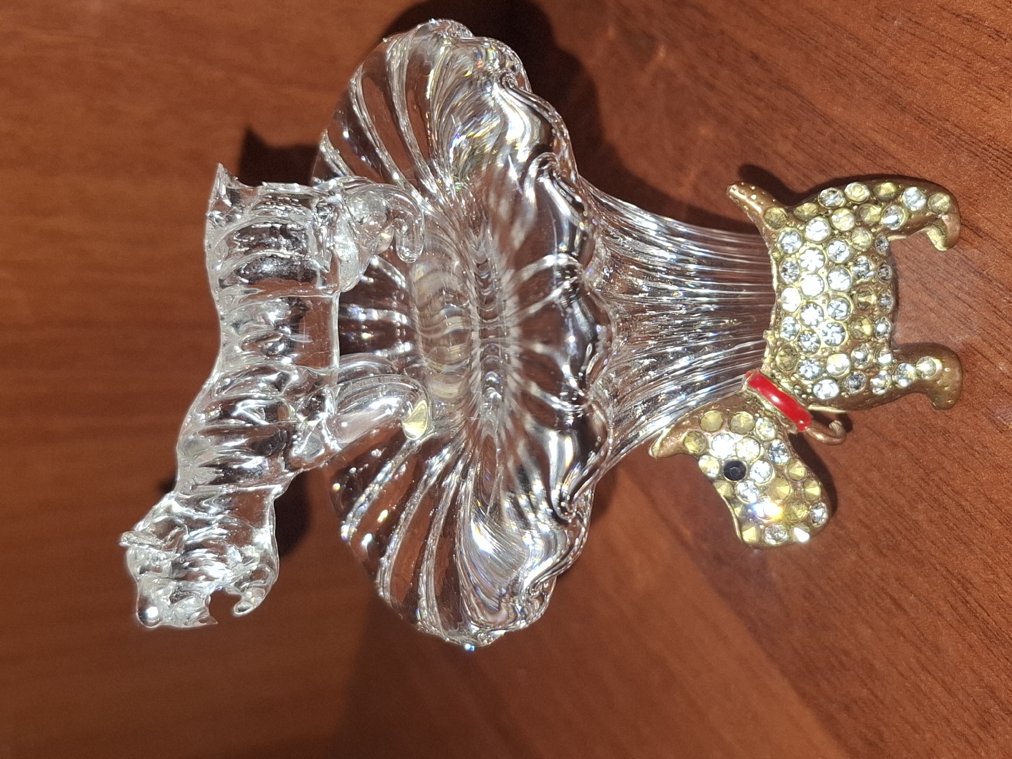 A close-up, eye-level shot shows a decorative figurine set. At the top, a clear glass tiger with its mouth open is perched on a clear glass pedestal. The pedestal has a fluted design, resembling a flower or a trumpet. Below the pedestal, a small, gold-toned dog figurine adorned with numerous clear and some dark rhinestones stands on a brown wooden surface. The dog wears a red collar. The background is a solid, warm brown color, possibly a wooden table or wall. The lighting creates highlights and reflections on the glass and rhinestones. This image depicts a decorative scene likely in a home or shop interior.