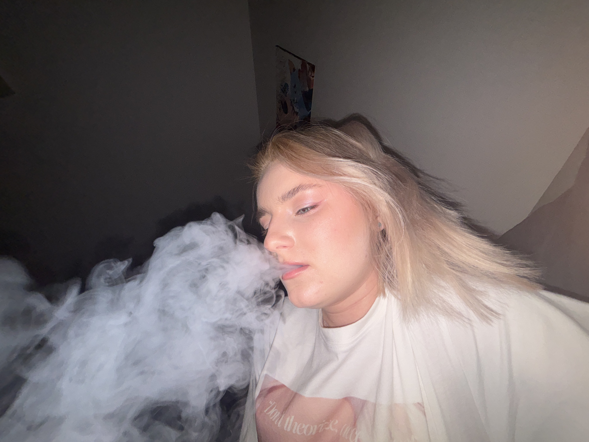 The image displays a single individual, appearing to be a young adult with blonde hair, lying on their back or side. The person is exhaling a large, dense cloud of white vapor or smoke from their mouth, which occupies the upper left portion of the frame. They wear a light-colored, possibly white, t-shirt with faint, curved text visible on the right shoulder/upper arm area. The individual has a fair complexion, with visible makeup including pink blush on the cheeks and subtle eye makeup. A thin necklace is visible around their neck. The setting is indoors and dimly lit, with a dark background transitioning to a lighter grey or tan wall on the right side of the frame. A blurred, rectangular object, possibly a framed picture or poster, is affixed to the wall in the upper right background. There are no discernible features or contextual clues within the image to suggest a specific geographical location, such as Ladozhskoe Ozero, Russia, or any other city or country.