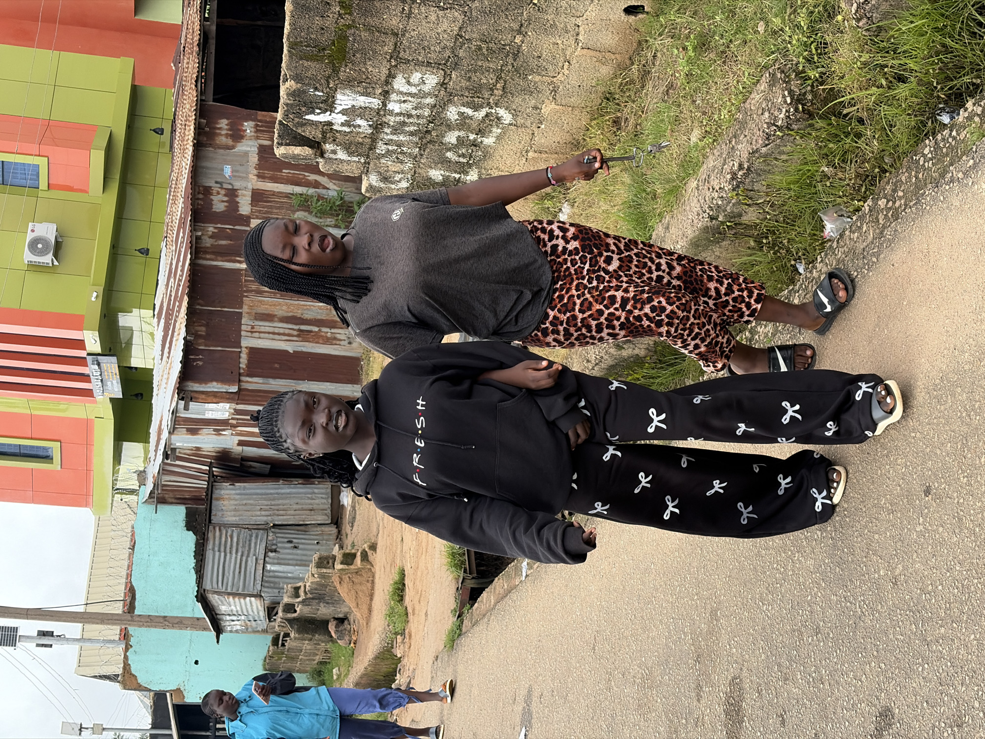 The image depicts two young women walking on a paved road in Jos, Nigeria. The woman on the left wears a black hoodie with "FRE·S·H" printed in white letters and black pants adorned with white bow patterns. The woman on the right is dressed in a grey t-shirt and a leopard-print skirt, and is holding a set of keys in her right hand. Behind them, a third person in a blue jacket walks away from the camera, holding a mobile phone. The environment is urban, characterized by a mix of modern and dilapidated structures. In the background, a vibrantly painted building with green and orange facades stands out, contrasting with adjacent corrugated metal shacks and a stone wall with white graffiti. The ground is a mixture of asphalt and dirt, with overgrown grass and a drainage ditch running alongside the road. The sky is overcast, suggesting a cloudy day.