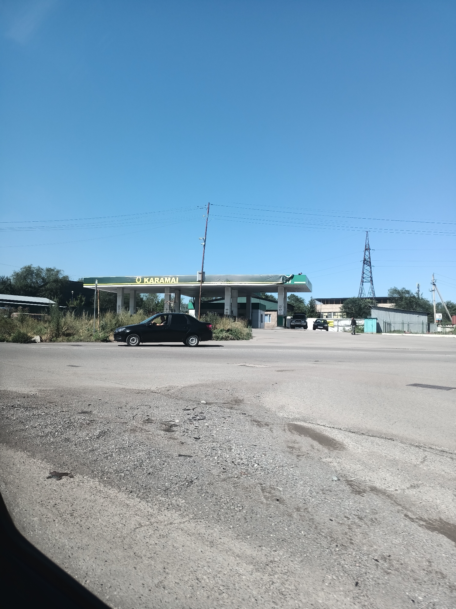 A black sedan is driving past a gas station with a green canopy and the logo "O KARAMAI". The gas station appears to be located in Talgar, Kazakhstan. The sky is clear and blue. The road is paved, and there are some bushes and trees in the background. There are also some other vehicles parked near the gas station and a tall transmission tower in the distance. The overall scene depicts a typical day at a gas station in a suburban or rural area.