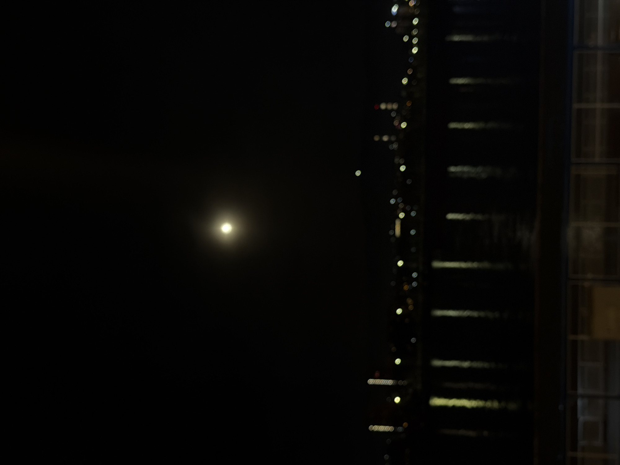 The image captures a nocturnal scene dominated by darkness. A prominent, bright circular light source, likely the moon or a distant artificial light, is positioned in the left-center of the frame, emitting a soft glow against the deep black sky. To the right, a vertical array of blurred, circular lights, typical of bokeh, extends along the edge, suggesting city lights or illuminated structures in the distance. Further to the right, a dark, vertical architectural element or building facade is partially visible. This structure features faint horizontal lines, possibly indicating windows or ledges, and a section with discernible rectangular panes, hinting at a building's exterior or a window frame. The overall impression is one of a quiet night in an urban or suburban environment, viewed from a slightly elevated position. There are no people or explicit activities depicted. The time of day is clearly night, with clear conditions allowing the bright light source to stand out. No text is visible in the image. Given the location context, this is a night view in Krasnoyarsk, Russia.