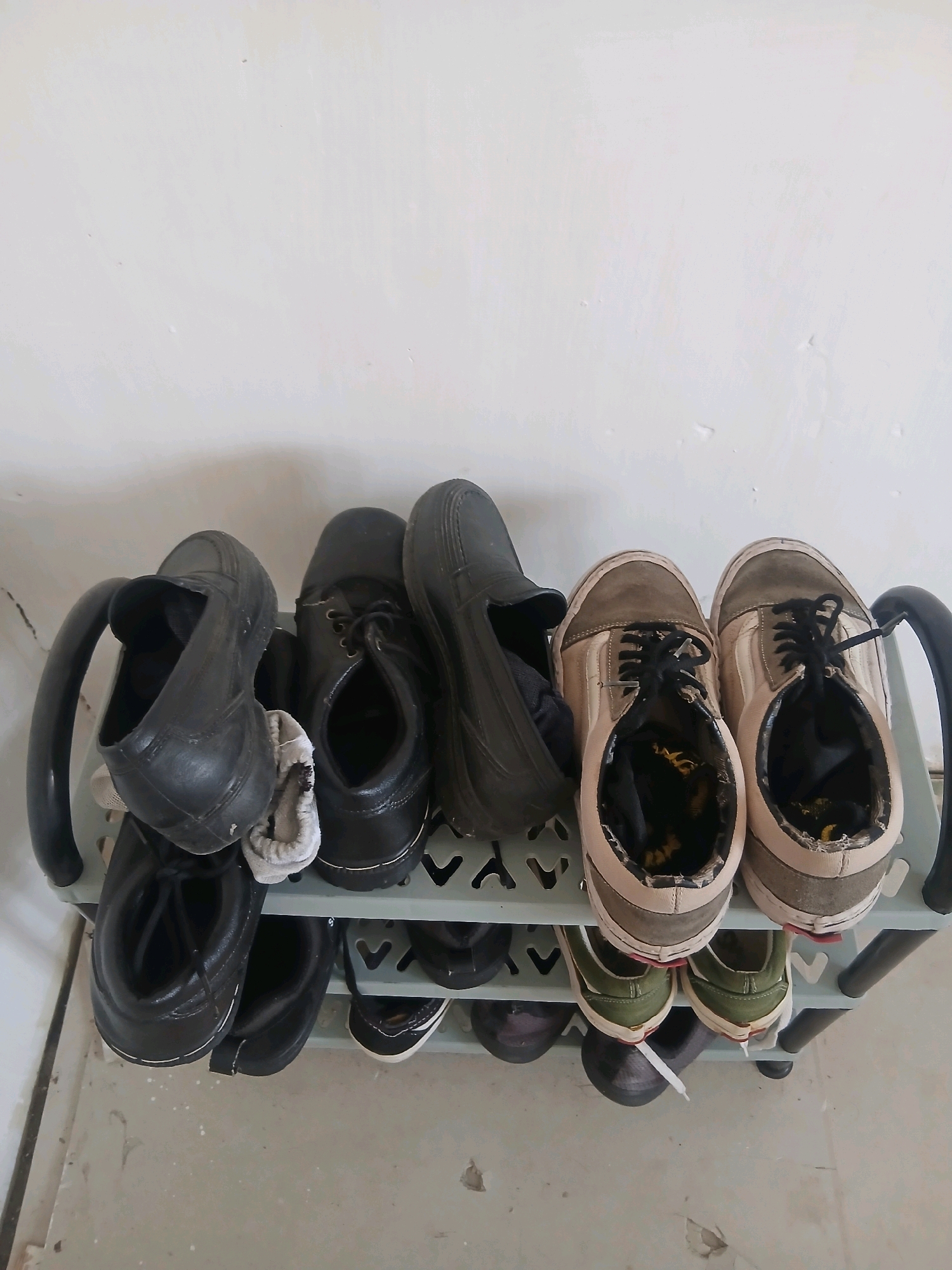 A shoe rack with multiple pairs of shoes is positioned against a white wall. The rack is made of gray plastic with black handles on each side. On the top shelf, from left to right, are two black slip-on shoes, a pair of black lace-up shoes, and a pair of tan and grey Vans. On the bottom shelf, from left to right, are two pairs of green and black sneakers, and two black slip-on shoes. The floor is concrete with some debris. The scene is indoors, possibly in Gorontalo, Indonesia, given the context.