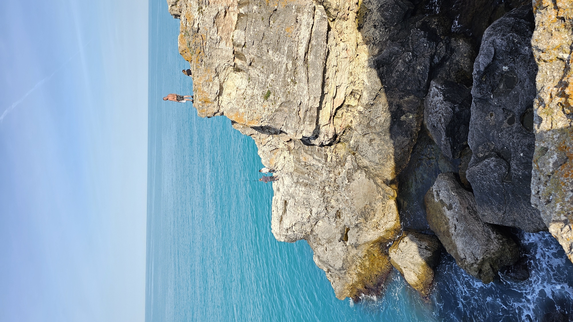 The image shows a coastal cliff scene. The water is turquoise and appears calm. A rocky cliff face dominates the right side of the frame, with various textures and colors suggesting different rock compositions. Several people are present on the cliff. One man stands at the edge, preparing to jump. Two others are visible nearby, watching or interacting. The location is in the city of Unknown, Unknown.