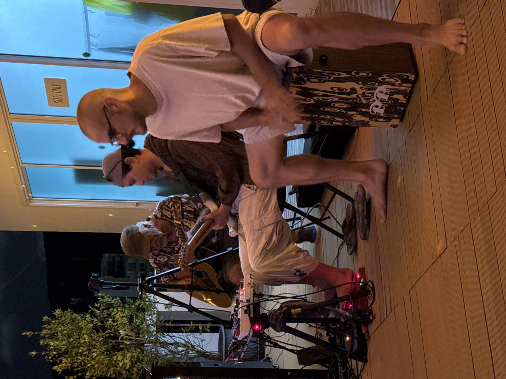 In Chalong, Thailand, three men are performing music outdoors on a wooden deck at night. The man on the left, wearing a patterned shirt and a cap, plays an acoustic guitar. In the center, a man in a brown hoodie also plays a guitar. To the right, a man in a white t-shirt and light shorts plays a cajón drum, his hands blurred in motion. Electronic music equipment is visible on a stand to the left. The background features a wall with large blue windows, one with a sign reading "STAFF ONLY." A lush green tree is positioned to the far left. The lighting suggests it is evening or night.