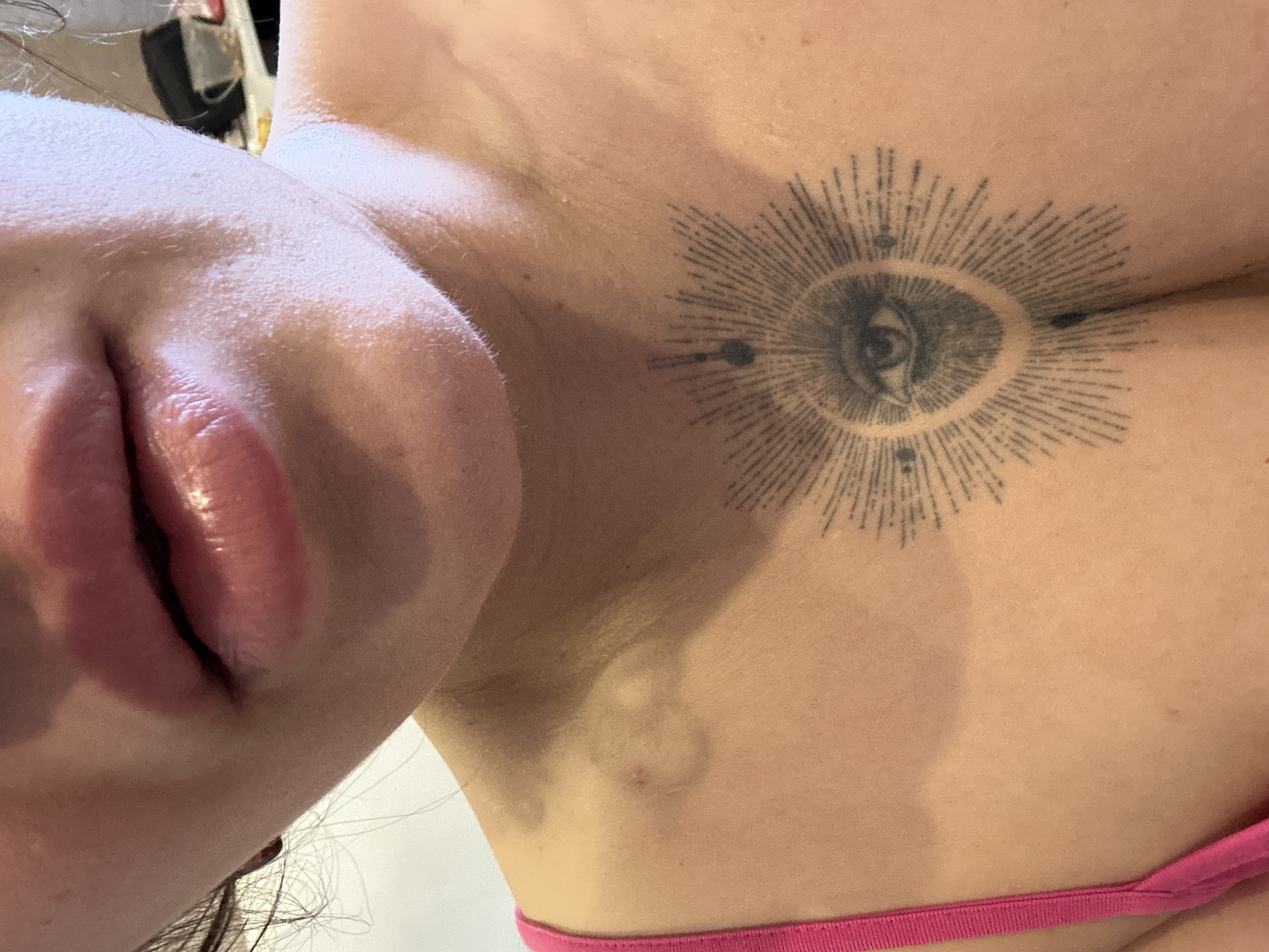 A close-up, slightly angled image captures the lower face and upper torso of an individual, presumed to be female. The full, glossy lips are slightly parted. The skin on the chin and surrounding the mouth exhibits fine vellus hair and visible pores. The neck and right shoulder are visible, with faint, circular indentations or marks present on the skin of the upper left shoulder/clavicle area. A bright pink strap of a brassiere or similar garment is visible along the left side of the frame. Centered on the upper chest, below the clavicle, is a dark-ink tattoo depicting a stylized "all-seeing eye" encased within a circular outline, surrounded by a radiant sunburst pattern. Small dots are symmetrically placed around the sunburst. The background is completely out of focus, indicating an indoor environment. The scene is situated in Chaikovsky, Russia.