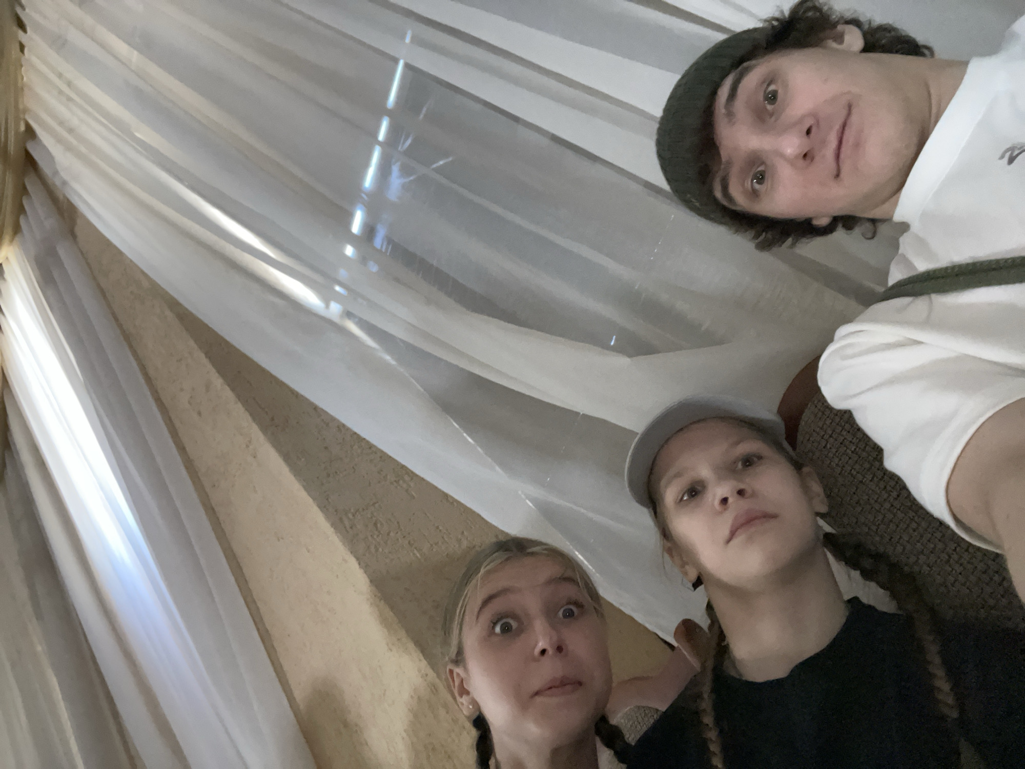 Three individuals are visible in this eye-level, upward-angled photograph. A male figure with curly brown hair wearing a dark green beanie and a white t-shirt is positioned in the upper right corner. Two younger individuals, both with blonde hair braided in pigtails, are in the lower half of the image. One is wearing a gray cap and a dark shirt, and the other is without a hat, wearing a light-colored top.
The backdrop consists of draped, translucent white curtains and a tan textured wall.
The location and city cannot be determined from the visual information provided.