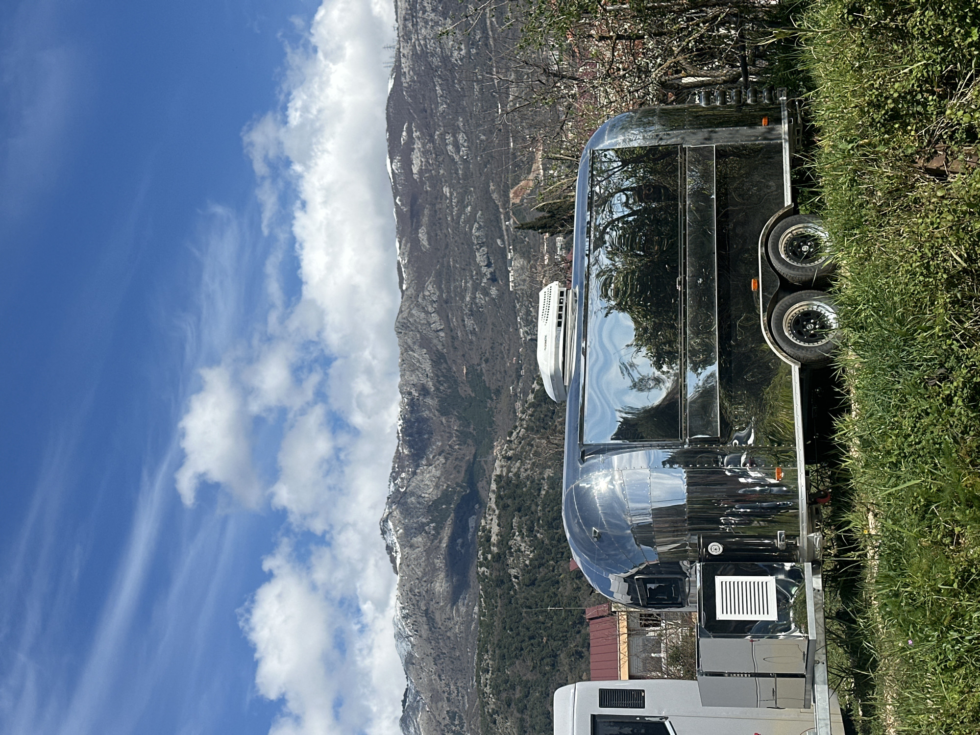 The image depicts a shiny, silver Airstream trailer parked near a mountain in Budva, Montenegro. The sky is blue with scattered white clouds. The Airstream is positioned on green grass. The front of a white RV is visible in the lower portion of the frame. A mountain range rises in the background.