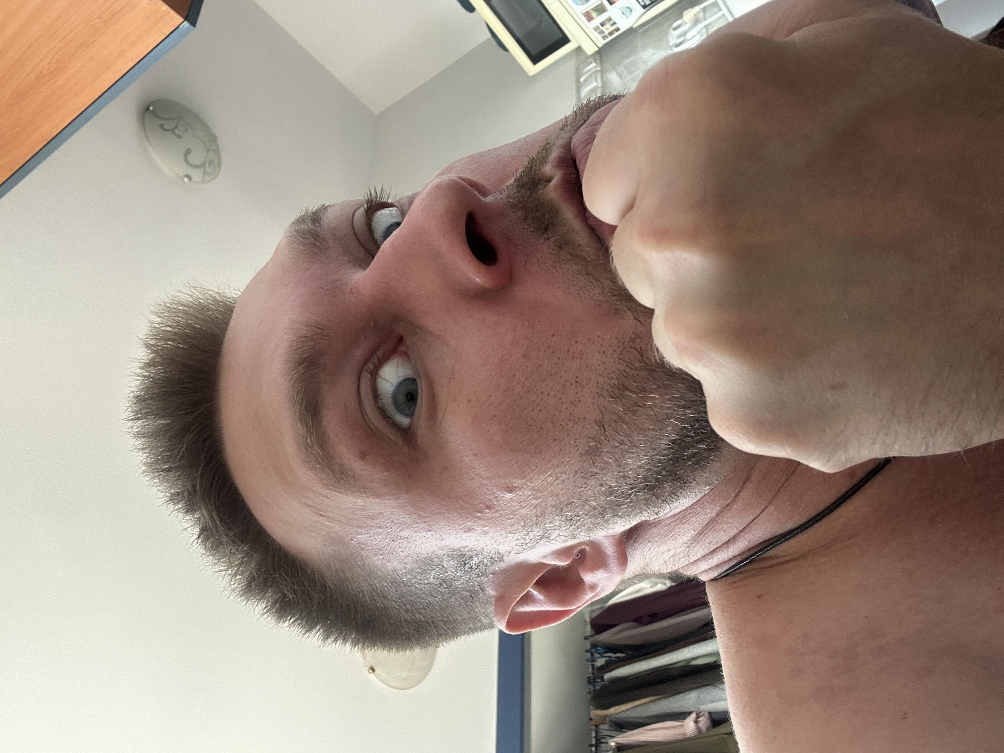 The media file shows a close-up, upside-down perspective of a person's face. The subject appears to be a light-skinned male with short, dark hair and stubble. His eyes are open and looking upwards, with one hand reaching towards his mouth. The background is blurred, but hints of an indoor setting are visible, including a wooden cabinet on the left and shelves filled with items on the right. The lighting suggests it is daytime. The scene is likely a casual, personal moment. There is no visible text in the image. The cropped images provide closer views of the subject's eye, nose, ear, and hand, as well as a decorative light fixture on the ceiling. The overall impression is an informal selfie taken from an unusual angle.