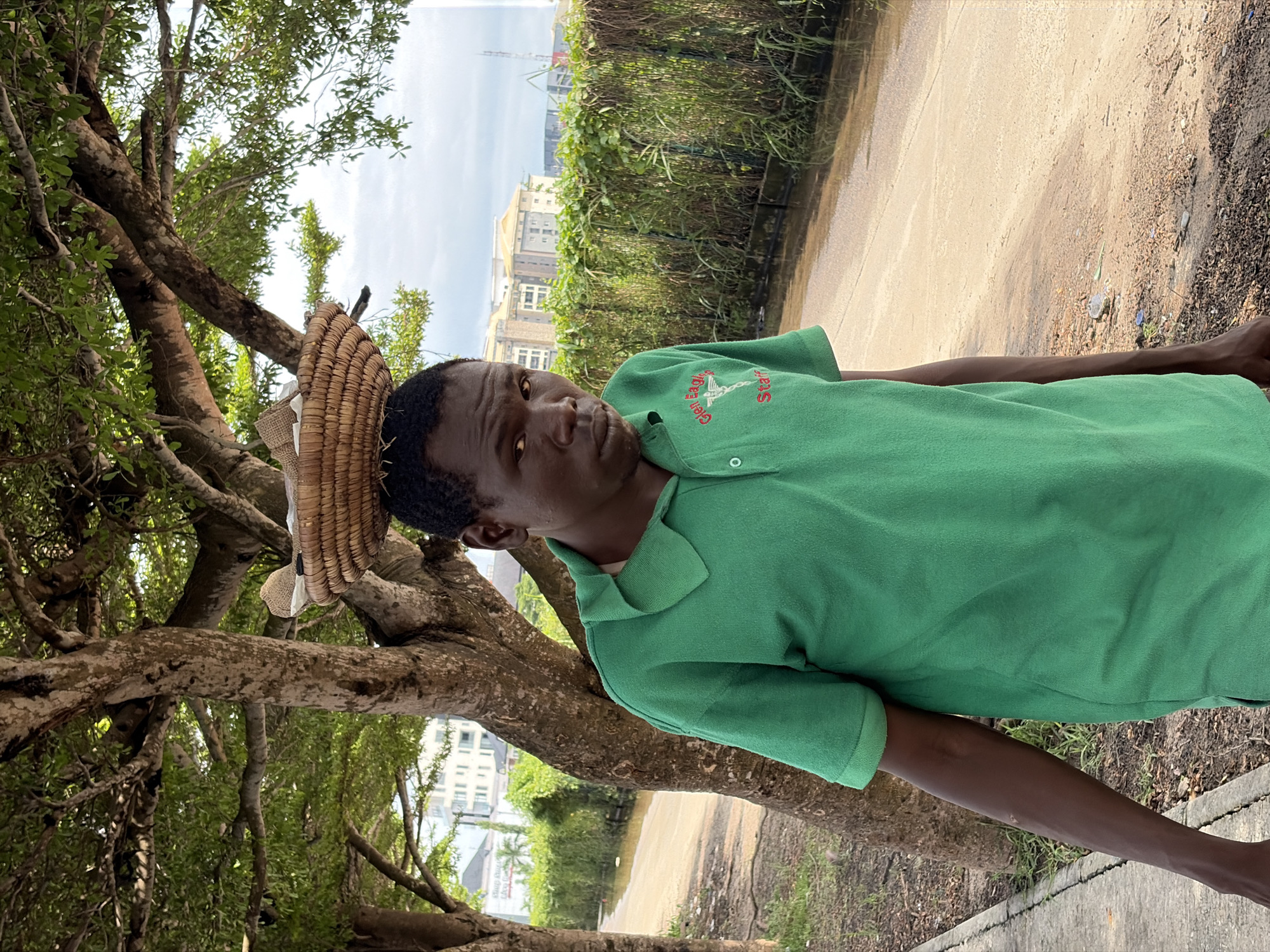 A medium shot captures a Black man wearing a green polo shirt, standing outdoors in Igboefon, Nigeria. He has short, dark hair and is looking directly at the camera with a neutral expression. A woven basket containing some items is balanced on top of his head. The man is standing next to a tree with a rough, textured trunk and branches covered in green leaves. Behind him, a dirt road runs alongside a flooded area with murky water. A hedge of tall, green grass and vines separates the road from a row of buildings in the background, which appear to be multi-story structures. The sky is overcast. The man's green shirt has an emblem on the left chest with red lettering that reads "Glen Eagles Staff".