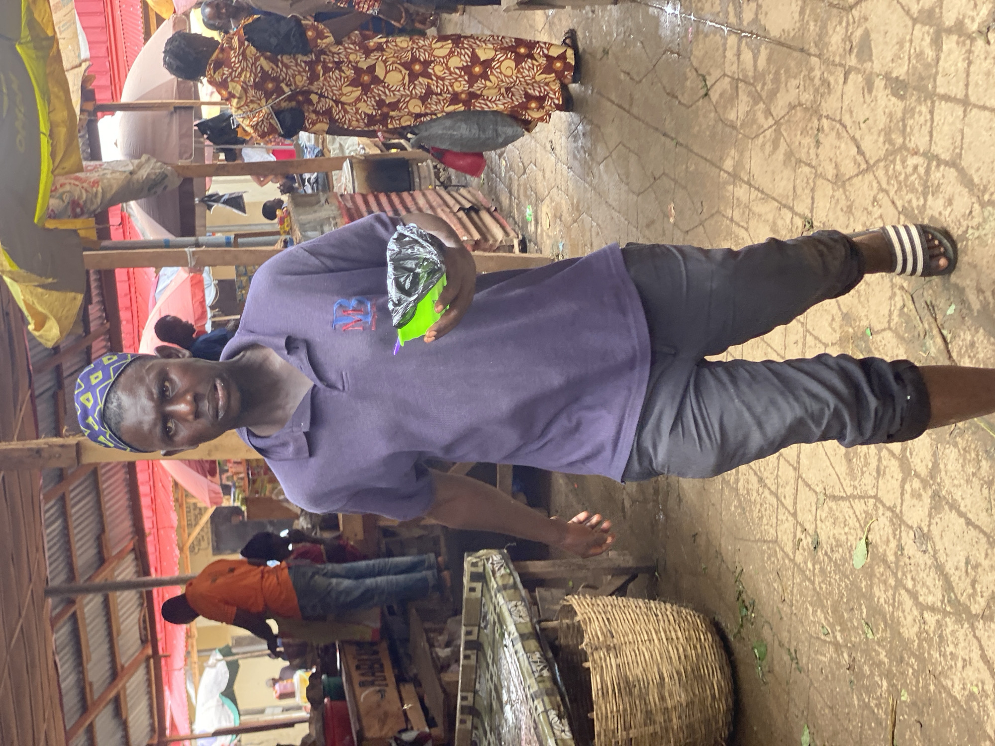 The image depicts a man standing in what appears to be an outdoor market in Kafanchan B, Nigeria. He is wearing a purple t-shirt with an emblem on the left side, dark pants, and striped sandals. He is holding a plastic bag containing a green substance.
The market setting includes stalls with corrugated metal roofs, various items for sale, and other people in the background. The ground appears to be a paved surface. The man is looking at the camera. There are people walking and selling items.