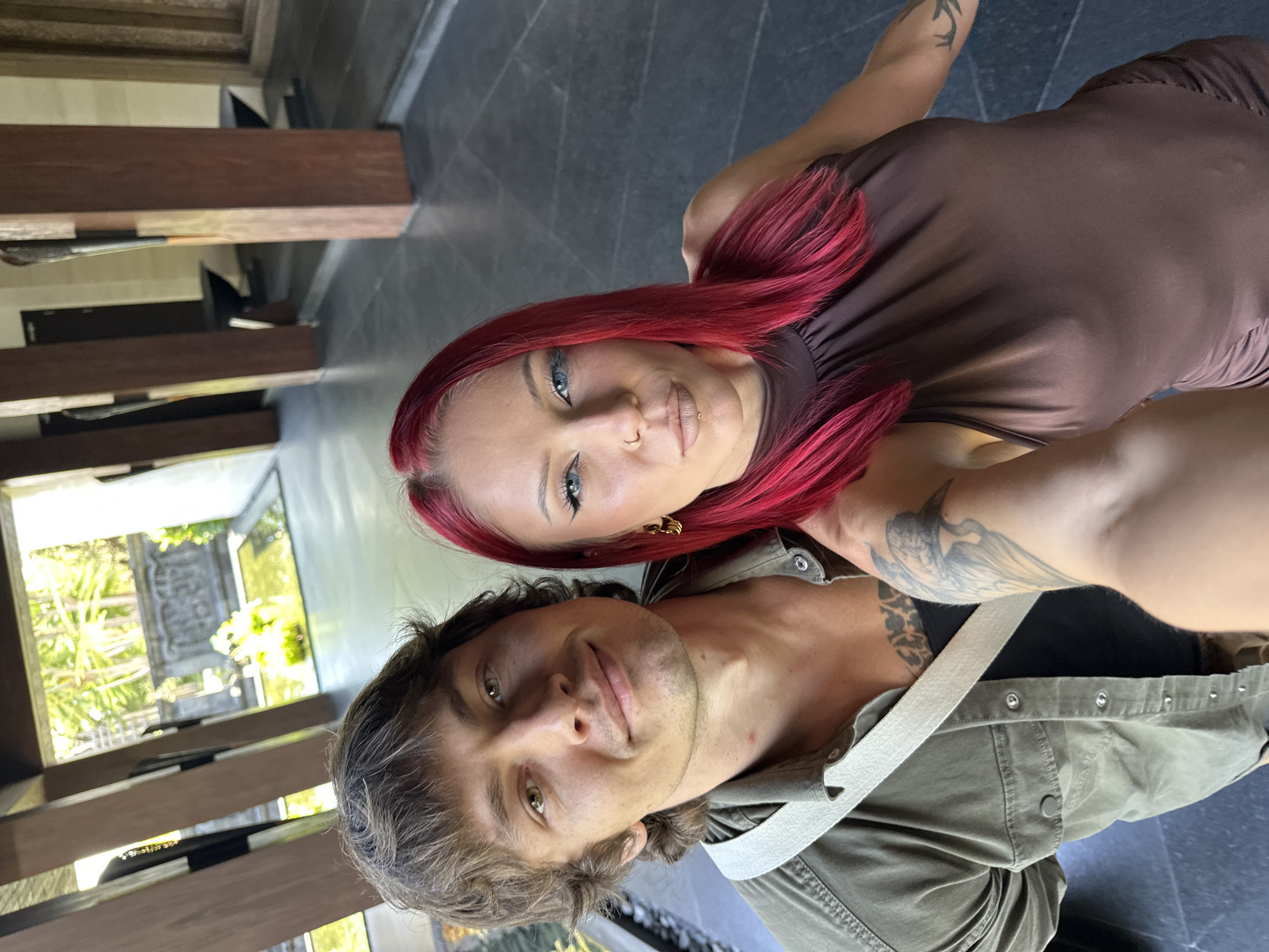 A selfie captures a man and a woman in Nusa Dua, Indonesia. The man, positioned at the bottom of the frame, has light brown hair and green eyes. He is wearing a green shirt with a white strap. The woman, situated above him, has red hair and blue eyes. She is wearing a brown dress. A tattoo is visible on her arm. The background features a blurred view of a building with wooden beams and greenery outside the window. The floor is tiled in dark grey.