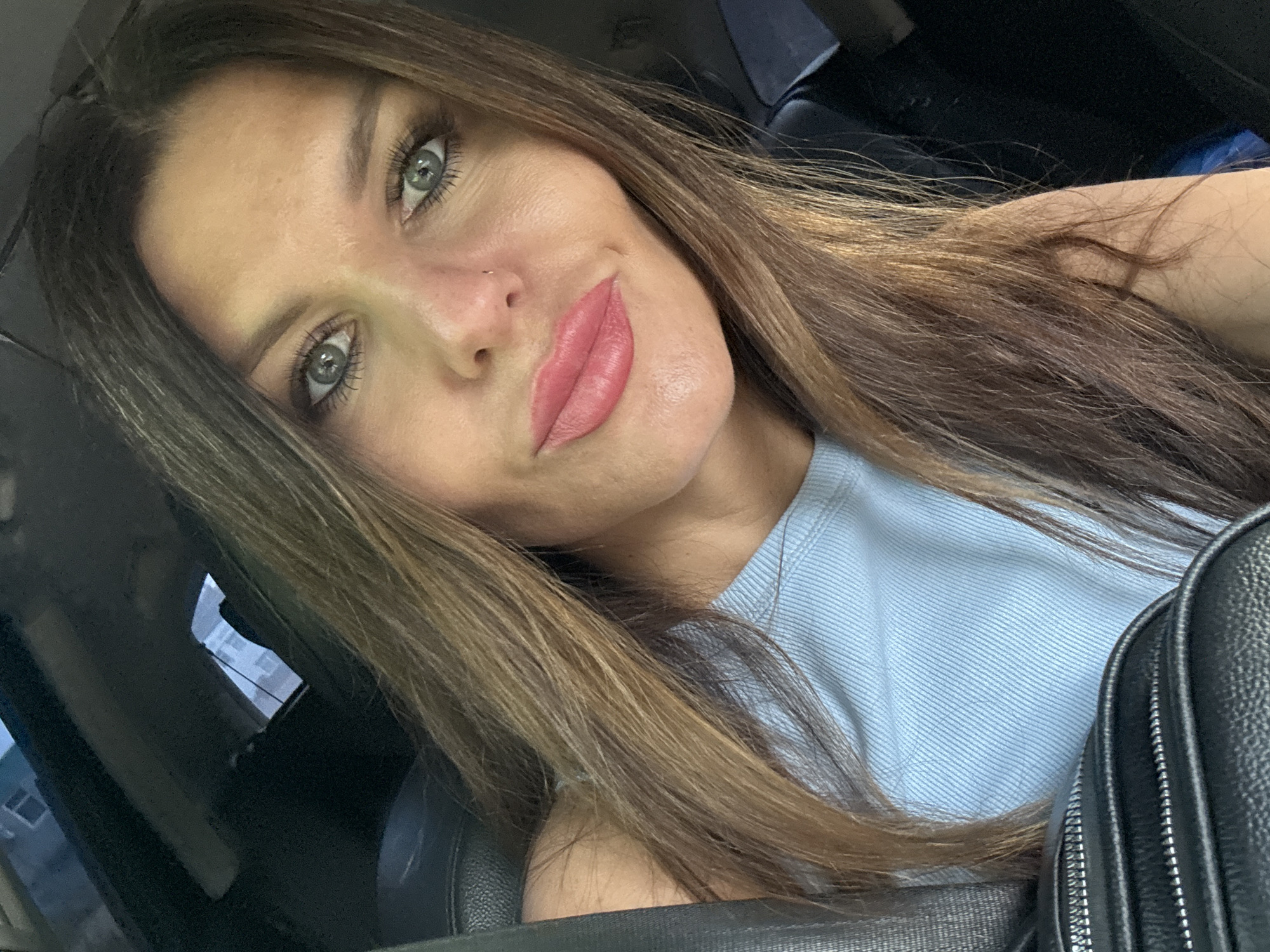 This is a close-up, selfie-style photo taken inside a vehicle. The subject is a woman with long, brown hair, green eyes, and pink lipstick. She is wearing a light blue ribbed top and a black bag is visible in the foreground. The background suggests an urban environment, with buildings visible through the car's windows. There is no specific indication that this image was taken in Chernushka, Russia.