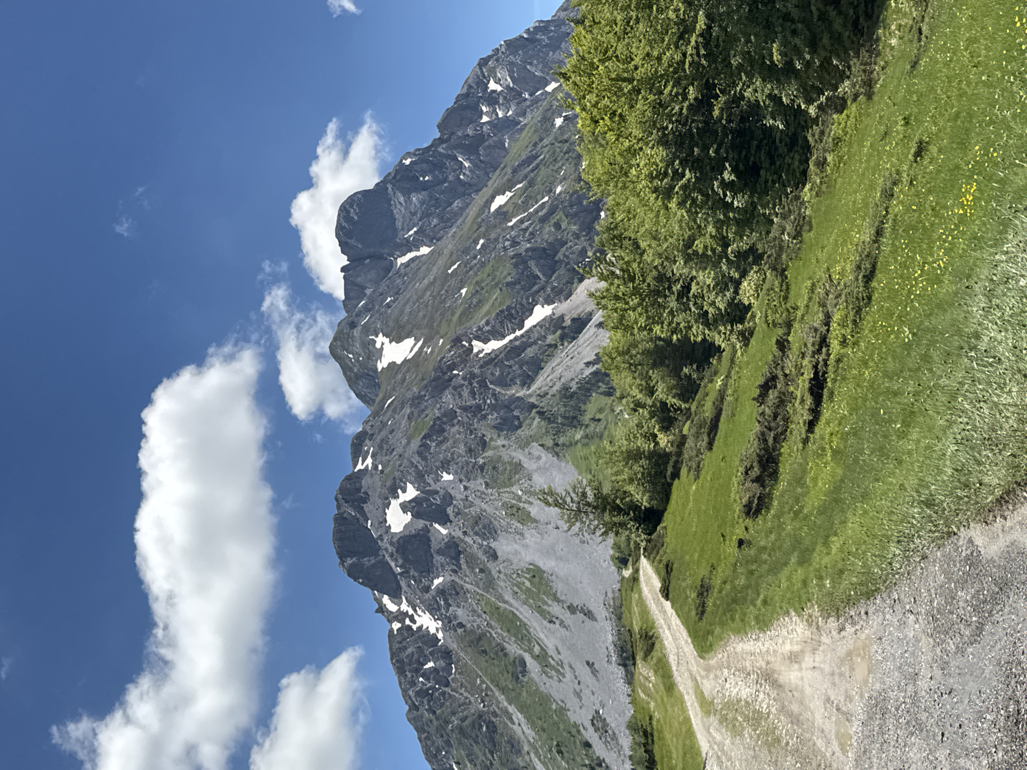 A dirt road winds up a grassy hillside towards a rugged, rocky mountain peak that is partially covered in snow. The mountain is dark gray and has steep, imposing slopes. The sky above is a clear, bright blue with scattered white, fluffy clouds. Lush green trees and vegetation cover the lower slopes of the mountain and the surrounding hillsides. The road appears to be the only man-made element in the natural landscape.