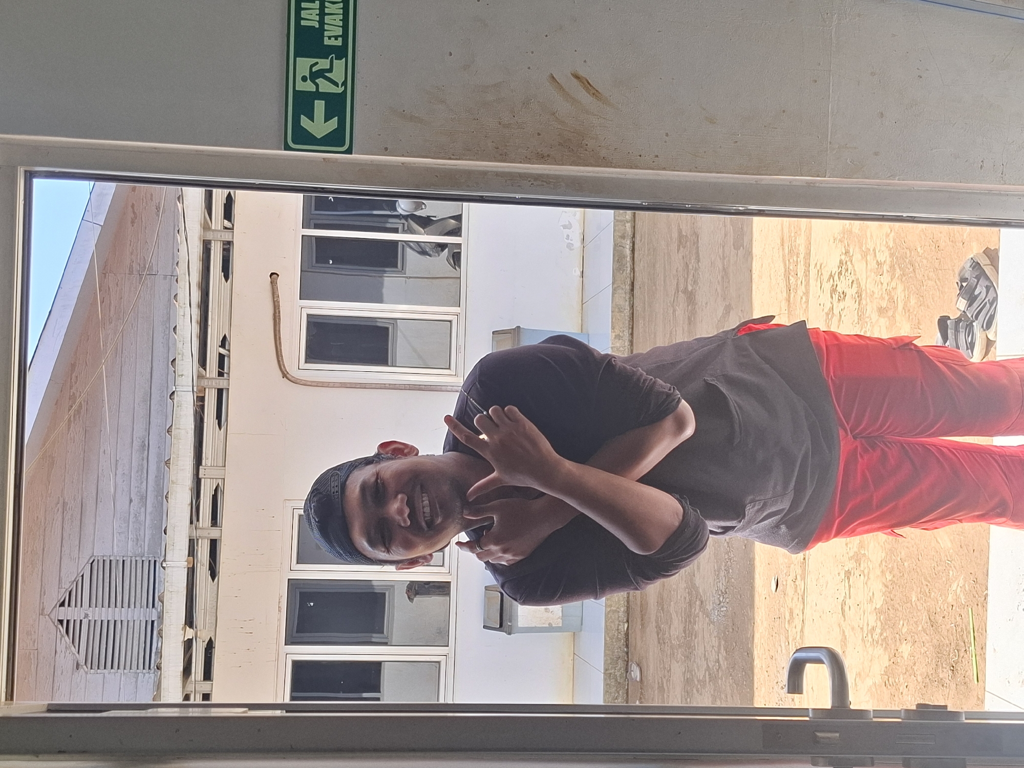 A young man wearing a black baseball cap, a dark long-sleeved shirt, and red pants stands in a doorway, smiling and crossing his arms in front of his chest. He is positioned in front of a white building with several windows and an exit sign on the right side of the frame. The ground outside is dirt and gravel. The scene appears to be in a non-urban outdoor setting.