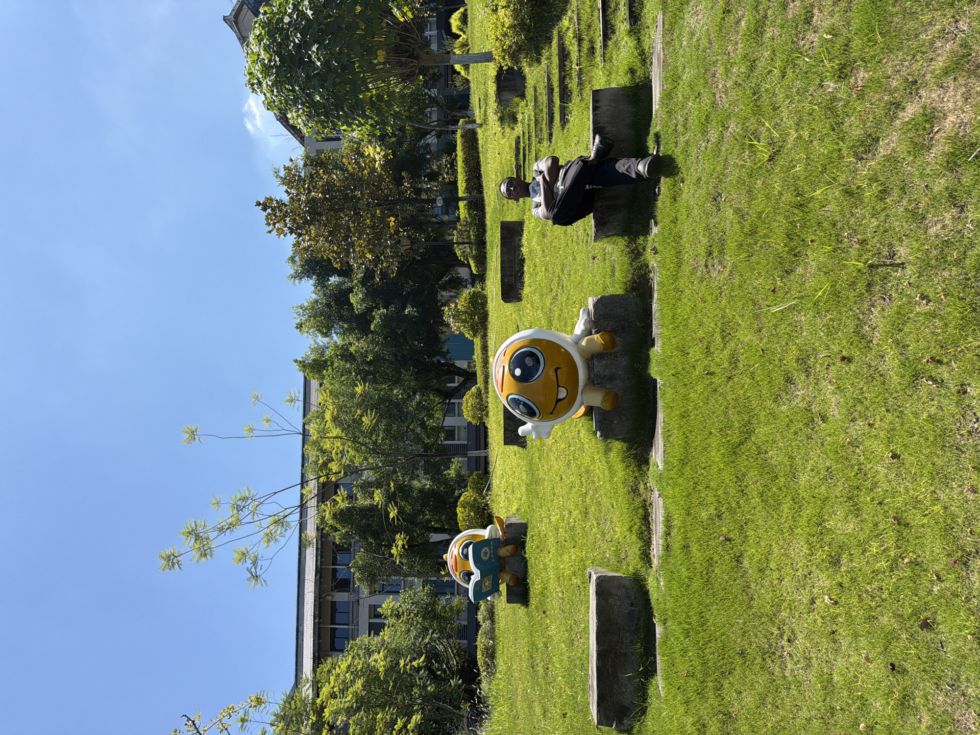 This image depicts an outdoor scene in the city of Jiangjin, China, on a sunny day with a clear blue sky. In the foreground, a lush green lawn is dotted with fallen leaves. Several concrete blocks are strategically placed on the grass. Two cartoonish, orange and yellow character statues are seated on these blocks. The statue on the left is depicted reading a blue book. The statue on the right is larger, with a wide smile and large eyes, and appears to be looking towards the viewer.
In the mid-ground, a person is seated on a concrete block to the right of the statues. The individual is wearing a dark shirt, black pants, and glasses, with arms crossed and legs crossed. They are looking towards the camera. Behind the statues and the person, there is a stepped pathway leading up a grassy slope.
Further in the background, a multi-story building with a gray roof and windows is visible, partially obscured by abundant green trees. The trees are varied in size and species, creating a shaded and natural environment. The overall impression is one of a serene and possibly educational or recreational outdoor space within a city park or campus.