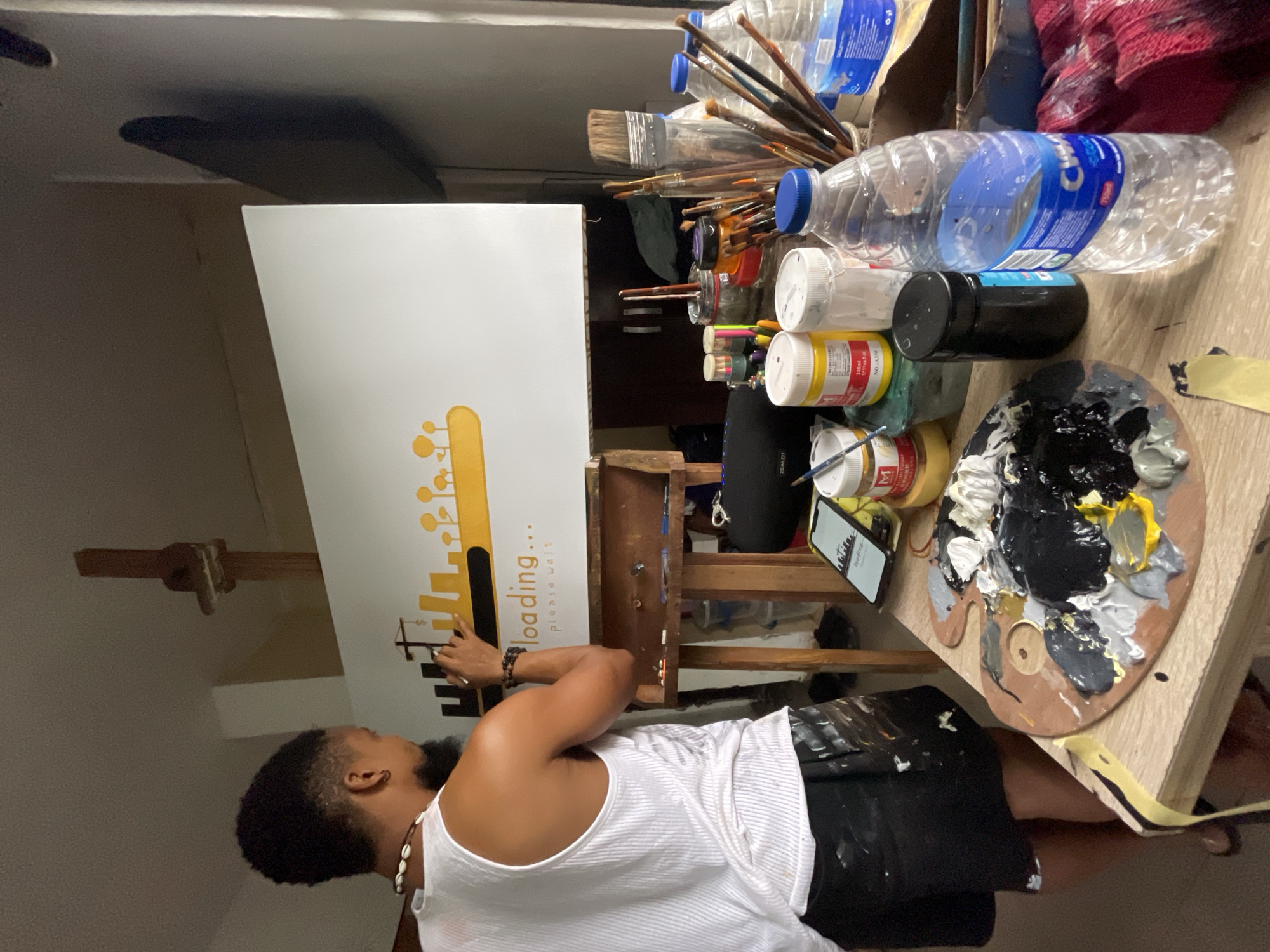 A man with dark skin and short hair stands in front of a canvas on an easel, seemingly painting. He is wearing a white tank top and black shorts. The canvas features a yellow graphic design depicting a stylized cityscape with a loading symbol, and the words "loading... please wait." The man holds a brush in his right hand and appears to be focused on his work.
An artist's workspace surrounds the easel, including bottles of water, paintbrushes, jars of paint, a black speaker, and a wooden palette with a mixture of black, white, gray, and yellow paints. A smartphone rests on the edge of the workspace near the palette.
The scene is indoors, and the location is indicated to be Ado, Nigeria. The lighting suggests an indoor environment, though specific details of the room are limited. A simple ceiling fixture can be seen.