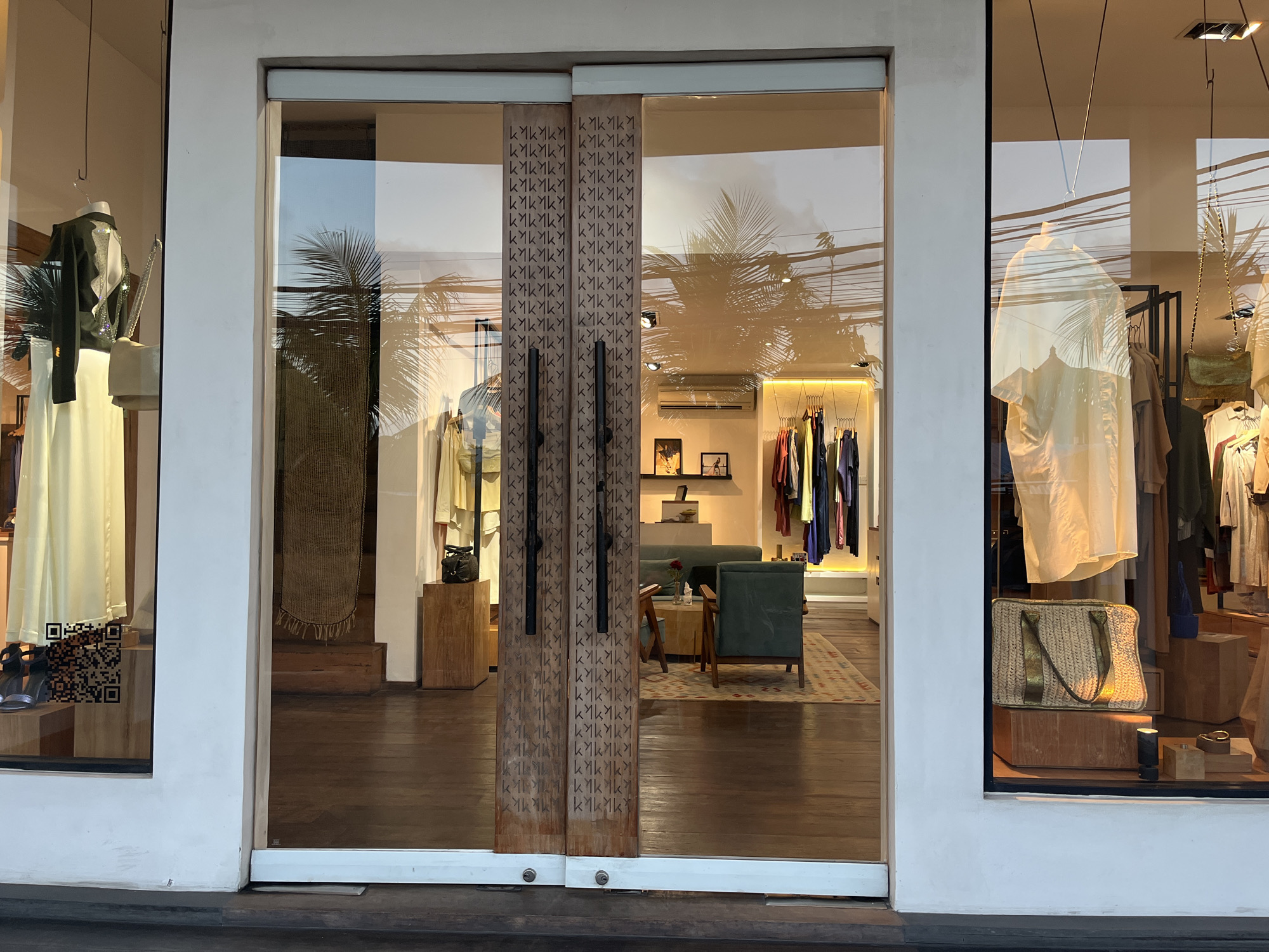 The image shows the entrance to a clothing store in North Kuta, Indonesia. The store has a large glass facade with double wooden doors in the center. The doors are decorated with a repeating pattern of what appears to be stylized letters or symbols. Through the glass, racks of clothing are visible, along with mannequins displaying outfits. There are also interior design elements such as sofas, tables, and rugs. Outside, palm trees are reflected in the glass, suggesting a tropical environment. The flooring in front of the store is dark wood.