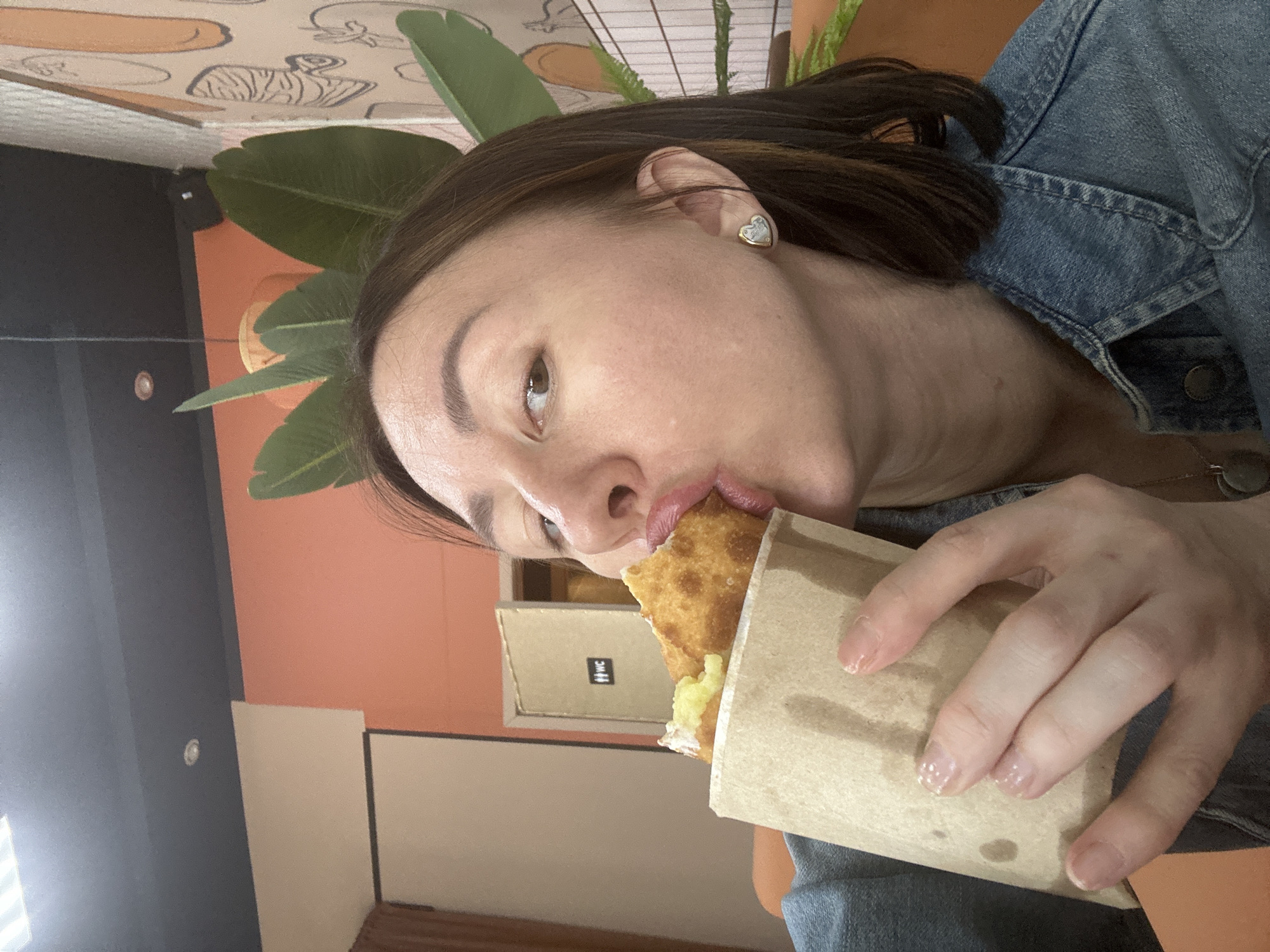 A woman with short brown hair and wearing a denim jacket is seen eating a pastry wrapped in brown paper. She is wearing a heart-shaped earring in her left ear. Behind her is a wall with orange paint and a decorative tropical plant. Also visible in the background is a sign with "WC" on it. This scene is located in the city of Votkinsk, Russia.