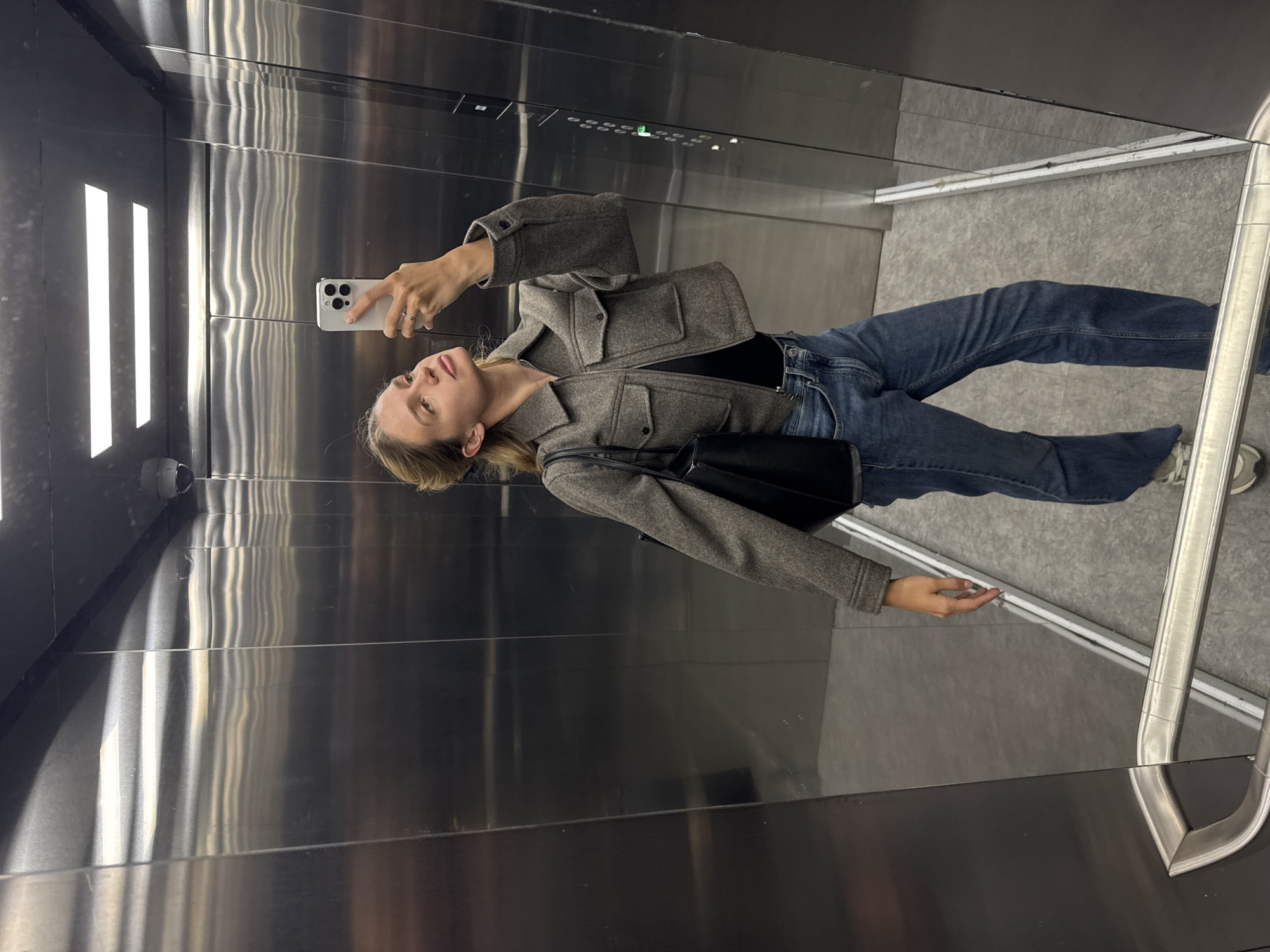 A young woman is taking a mirror selfie inside an elevator. She is positioned slightly to the left of the frame, facing the camera reflected in the polished stainless steel wall. She holds a light-colored iPhone, presumably white or silver, with her left hand, which has a ring on the index finger. Her right hand is relaxed at her side.
She is wearing a grey, textured, collared jacket with pockets on the chest, a black top underneath, and blue jeans. Her hair is light-colored and appears to be pulled back or short, revealing her ear. She has a neutral or slightly contemplative expression, looking directly at the phone's camera. A black shoulder bag hangs over her right shoulder.
The elevator interior is predominantly clad in reflective stainless steel panels, showing streaks and reflections of the internal lighting and the woman. Bright vertical strip lights are strongly reflected on the left wall. The floor appears to be a light grey, possibly tiled or concrete, and a silver metal handrail extends along the right side of the elevator, reflecting the person's sneakers. On the upper wall, above the woman's head, small green illuminated indicators or buttons are visible, suggesting the elevator control panel. There is no clearly visible text on any part of the scene or the subject's clothing. The overall lighting is bright, illuminating the reflective surfaces of the elevator.