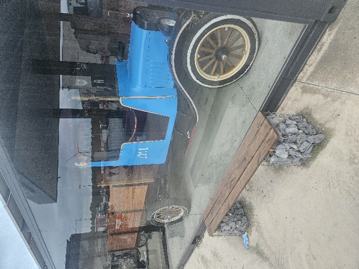 The image displays a vintage utility vehicle exhibited behind a large glass panel, located in Tudela, Spain. The vehicle features a bright blue body, dark fenders, and an open top with a light-colored wooden cargo bed at the rear. It possesses white-sidewall tires and multi-spoke wooden wheels. A white stylized 'Nº1' marking is visible on the blue side panel.
The glass enclosure reflects an overcast sky, a distant white wind turbine, other vehicles resembling trucks, indistinct human figures, and partial text including 'HUMANCO' and a crest emblem, indicating an open commercial or industrial area.
In front of the glass, on a concrete surface, a bench is present. The bench consists of a wooden plank seat supported by two rectangular gabion baskets filled with grey stones. Small patches of green vegetation are visible around the gabion bases, along with a piece of blue plastic debris.
