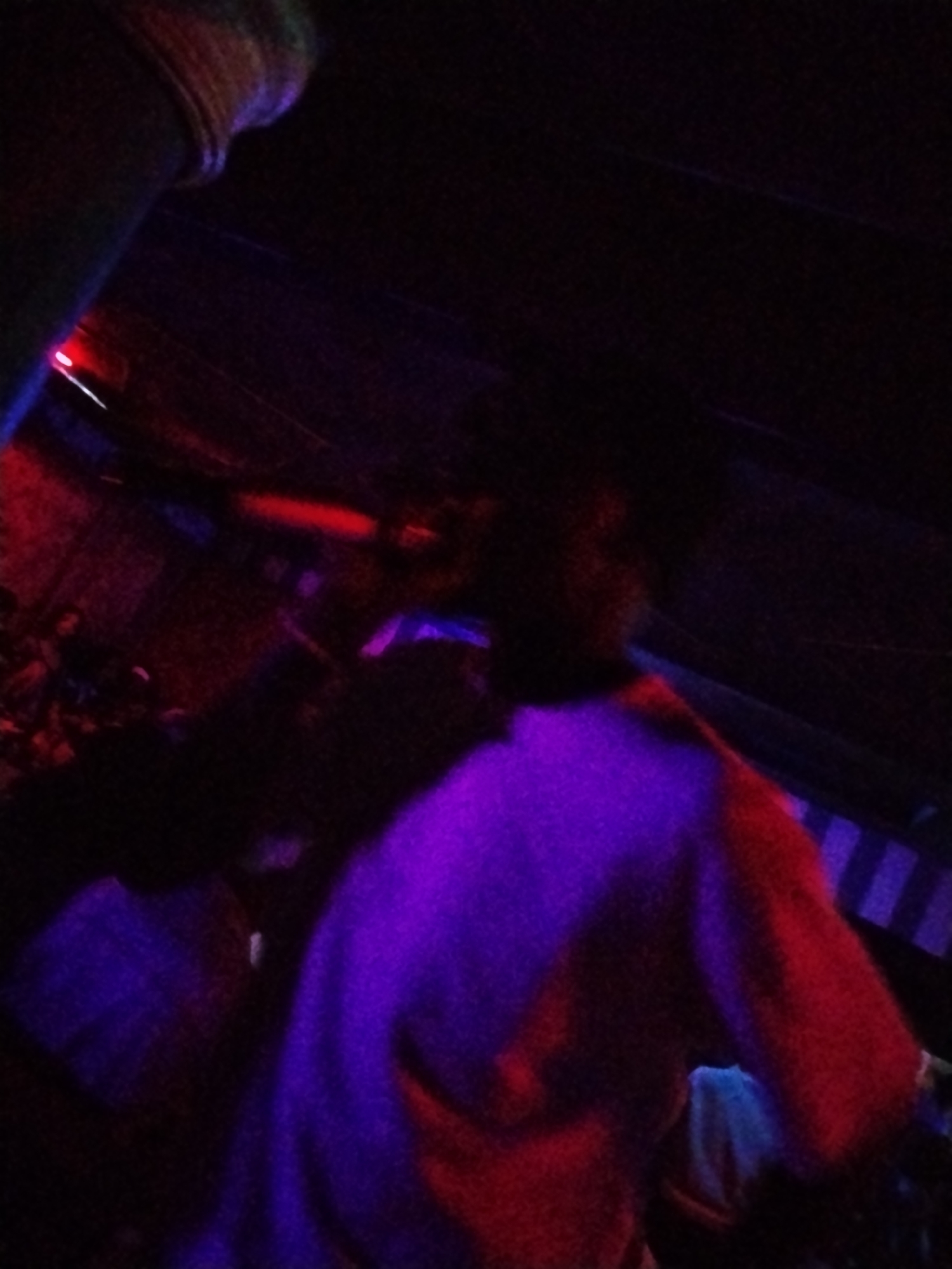 This is a poorly lit, blurry image taken indoors at night, likely in a venue such as a club or concert hall in Durumi, Nigeria. The scene is dominated by dark shadows and vibrant, artificial lighting, primarily purple and red. In the foreground, a person is visible, partially illuminated by a bright purple light reflecting off their clothing, which appears to be a white or light-colored top or jacket. The person's head and upper body are captured, and their gaze is directed downwards or away from the camera. In the background, other individuals are present, but they are indistinct due to the low light and motion blur. The environment appears to be a crowded space with some structural elements visible, such as what looks like a pipe or beam in the upper left corner. The overall atmosphere suggests an event with dynamic lighting and active participants.